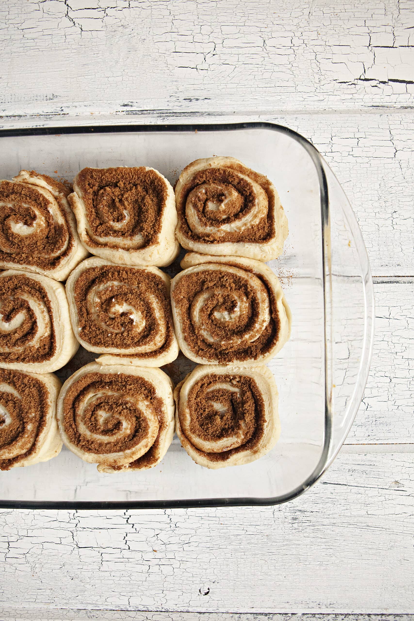 Uncooked cinnamon rolls in a glass baking dish on a white wood background.