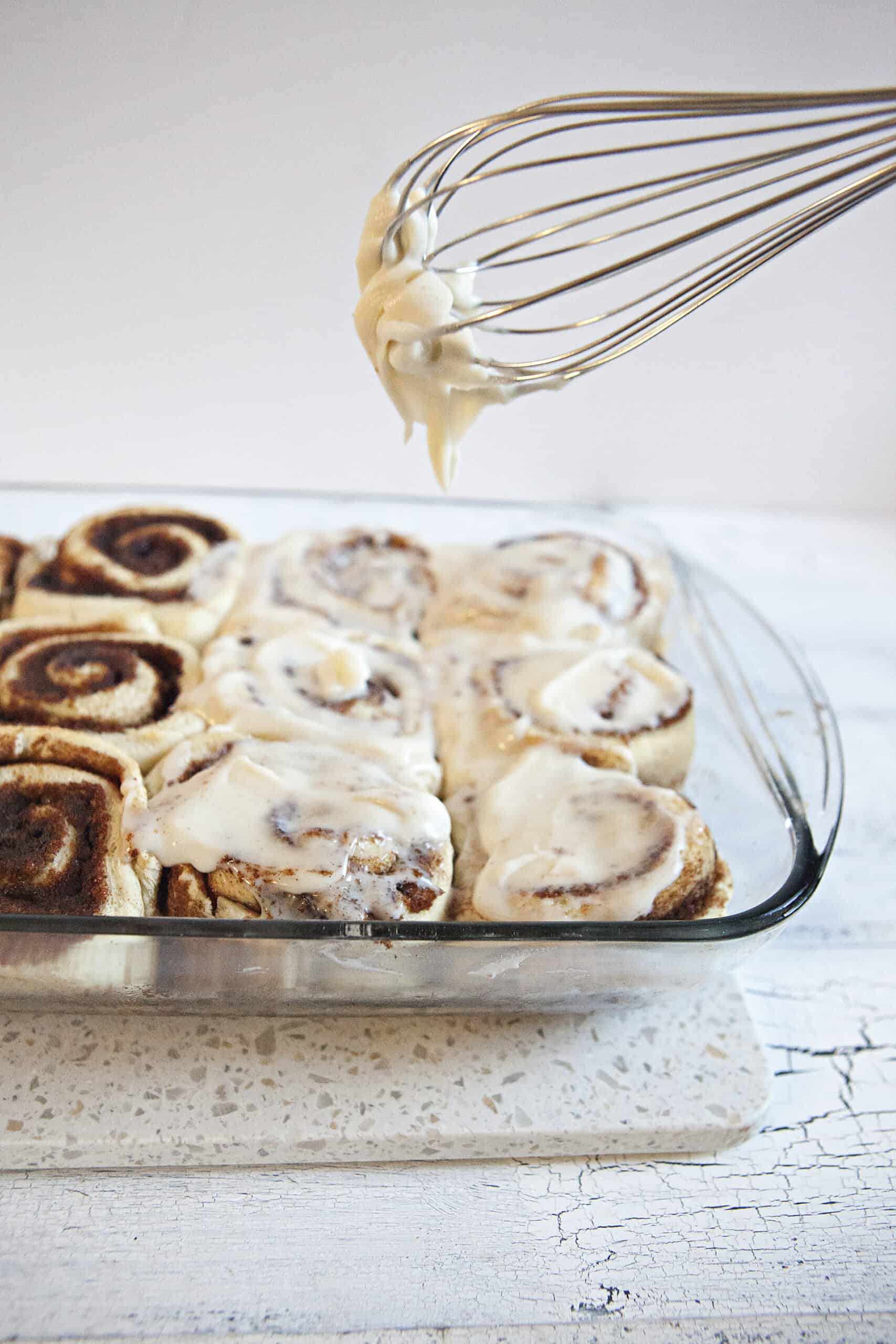 Soft and fluffy cinnamon rolls in a baking dish, half of the rolls topped with cream cheese frosting, with a whisk holding more frosting dripping onto the rolls from above.