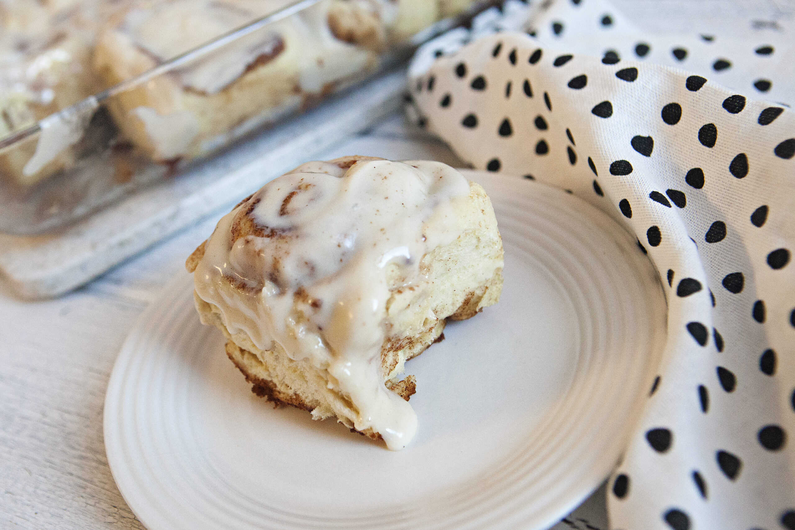 Soft and fluffy cinnamon roll with cream cheese frosting on a white plate next to a white napkin with black polka dots with a baking dish of cinnamon rolls in the background.