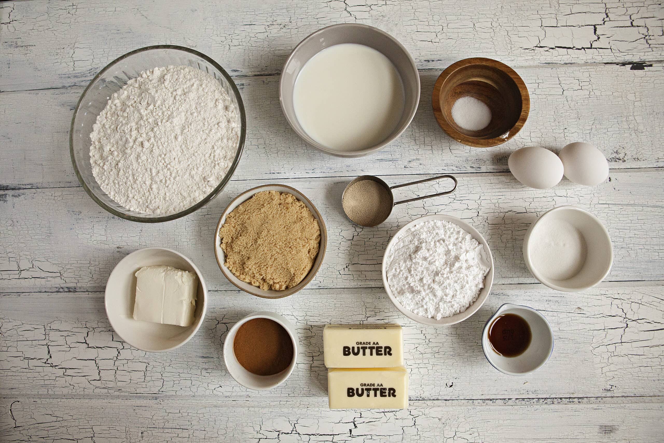 Ingredients for soft and fluffy cinnamon rolls with cream cheese frosting:  Flour, milk, sugar, salt, brown sugar, eggs, yeast, cinnamon, butter, cream cheese, powdered sugar, and vanilla, on a white wood background.