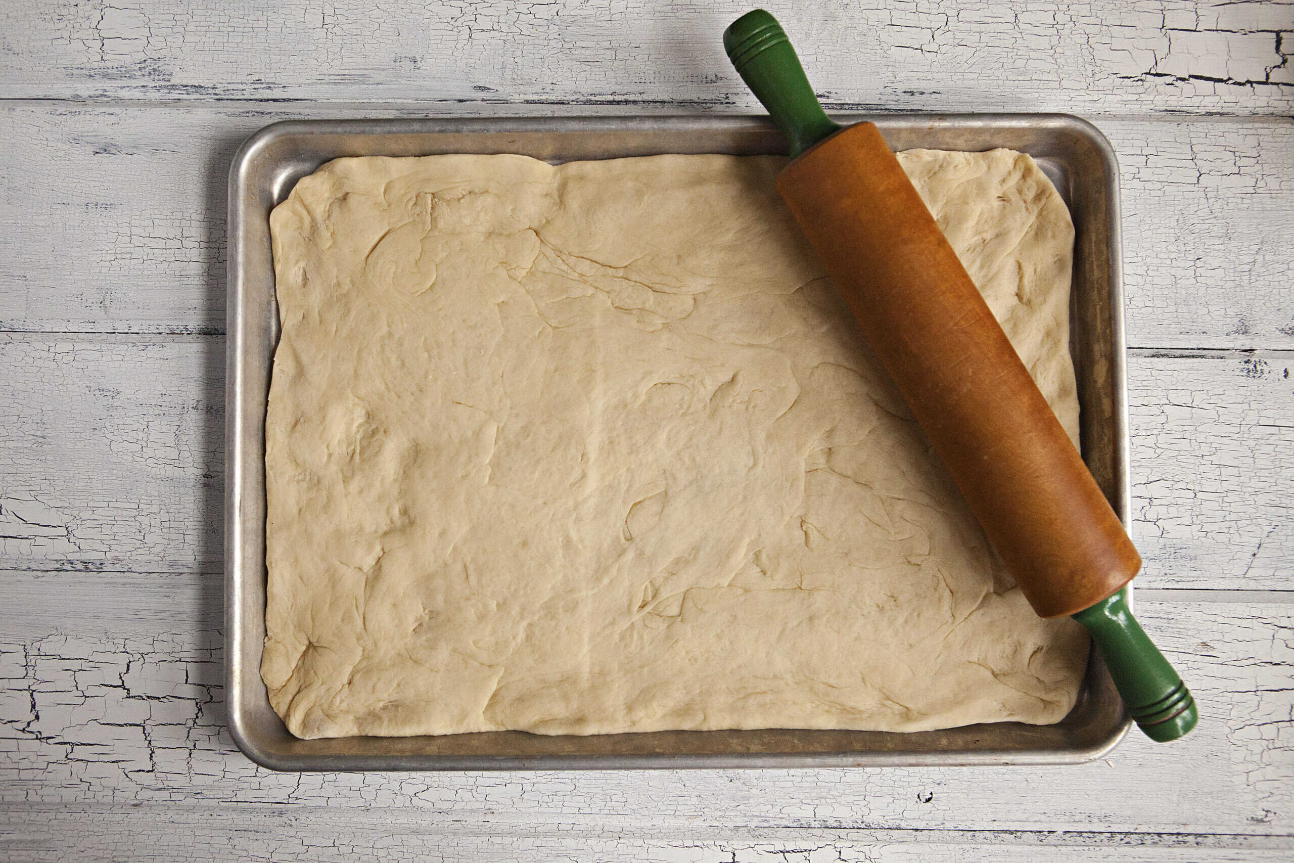 Dough for soft and fluffy cinnamon rolls being rolled out on a baking sheet with a rolling pin on a white wood background.