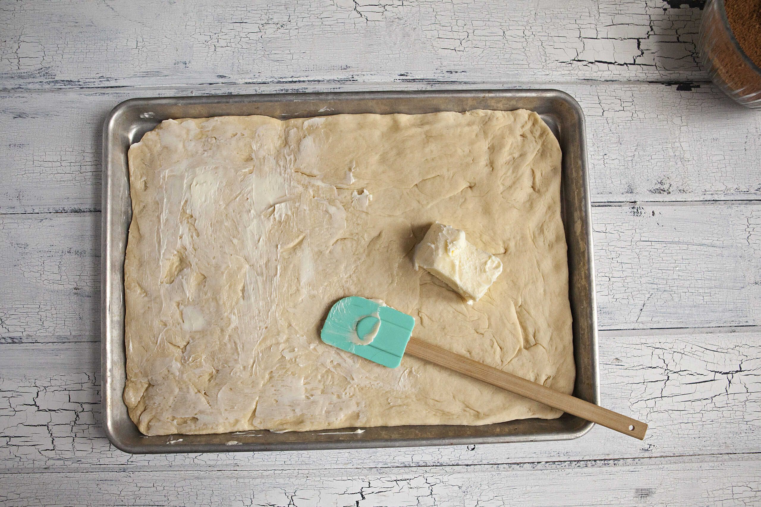 Dough for soft and pillowy cinnamon rolls rolled out on a baking sheet, being covered with softened butter with a blue spatula, on a white wood background.