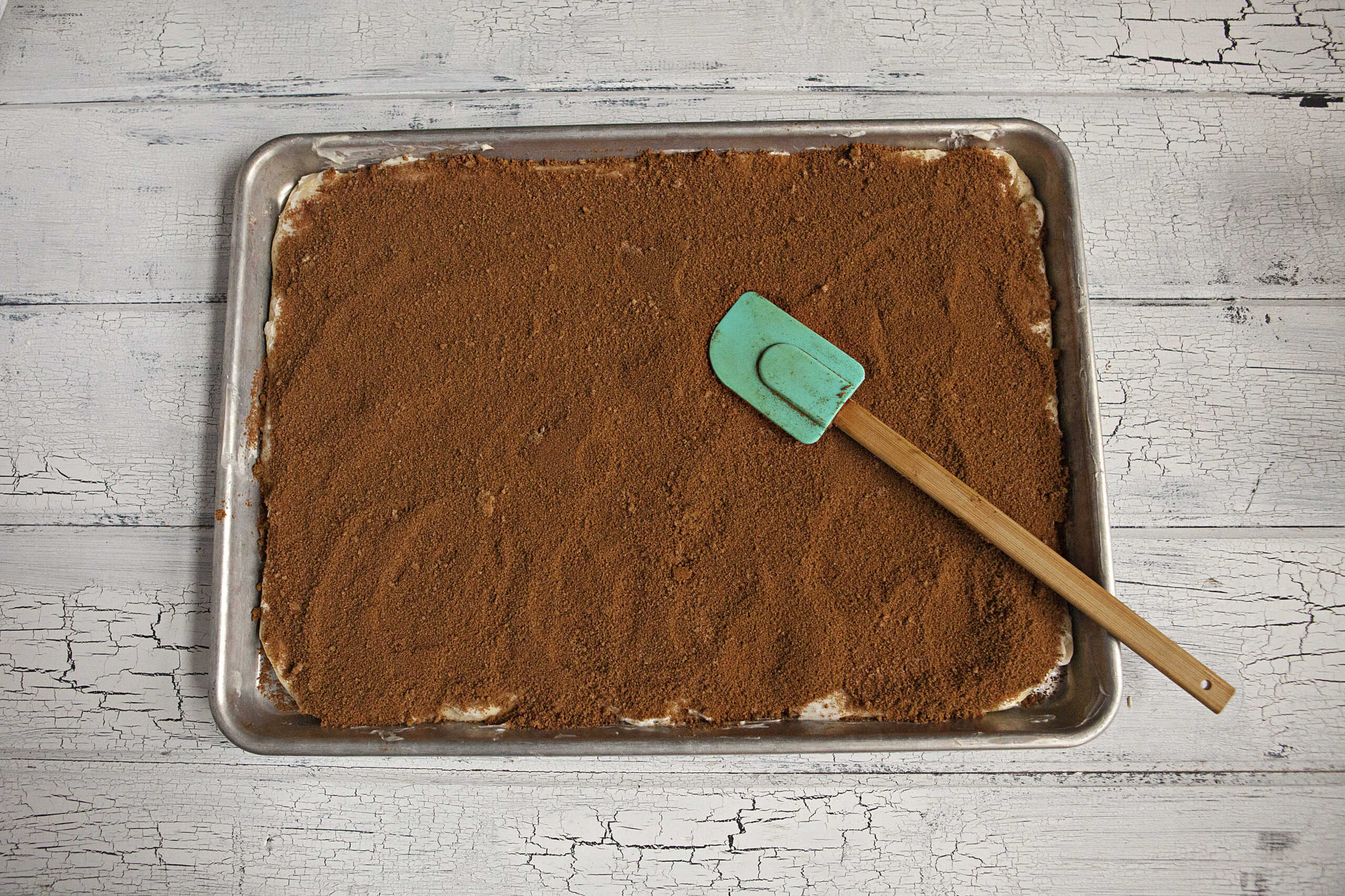 Dough rolled out for soft and pillowy cinnamon rolls on a baking sheet being covered with a cinnamon/brown sugar mixture by a blue spatula on a white wood background.