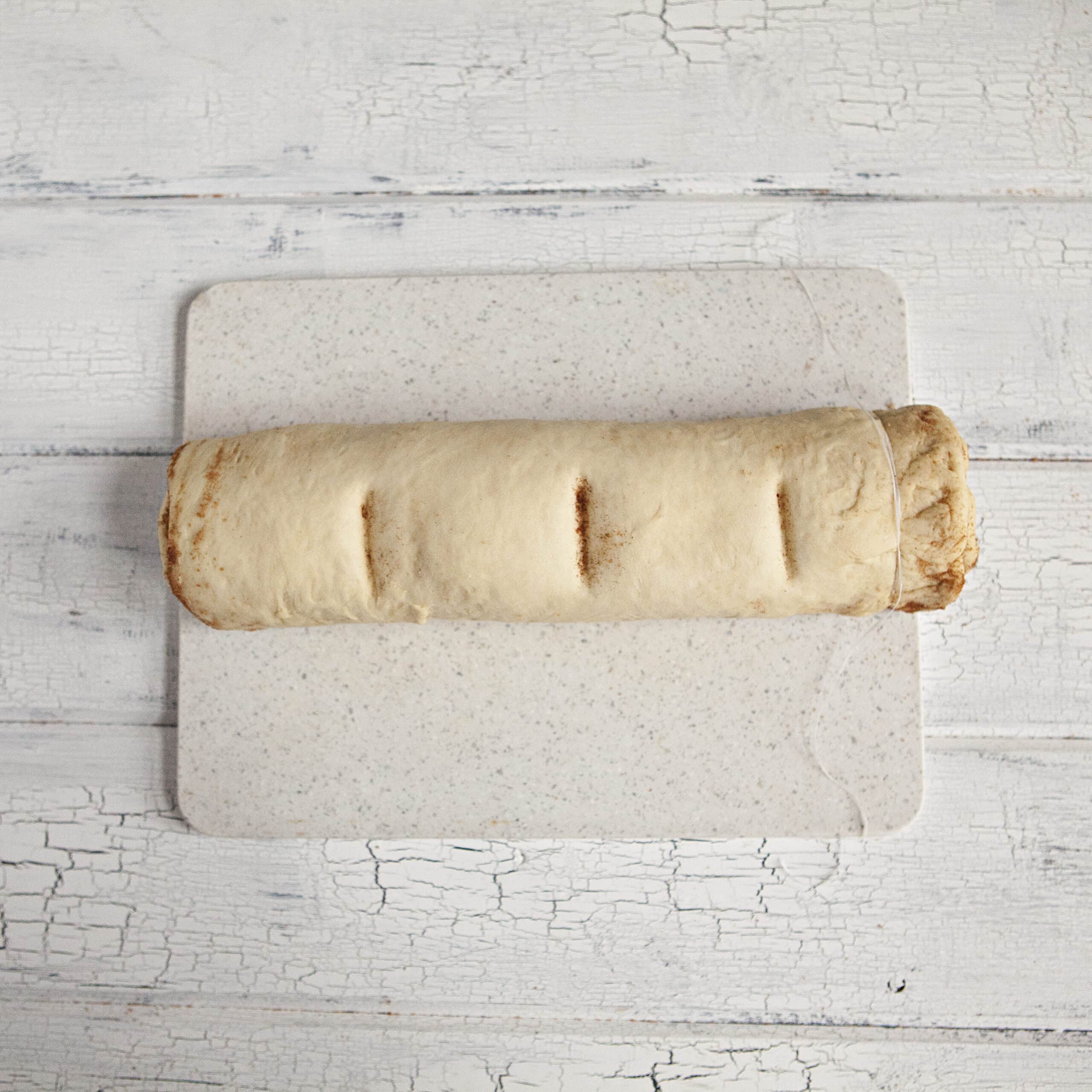 Dough for soft and fluffy cinnamon rolls rolled into a cylinder with indentations to mark the place for cutting the cinnamon rolls into equal portions, on a white cutting board and a white wood background.