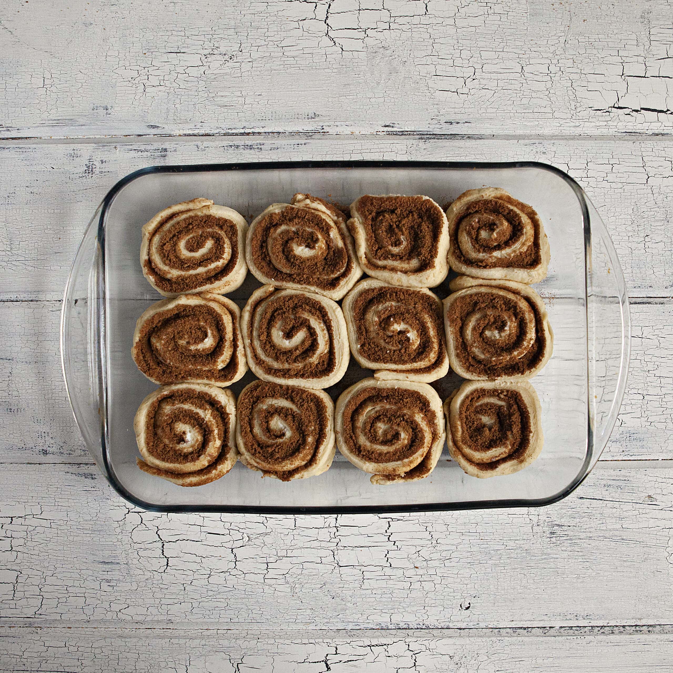 Uncooked soft and fluffy cinnamon rolls in a glass baking dish on a white wood background.