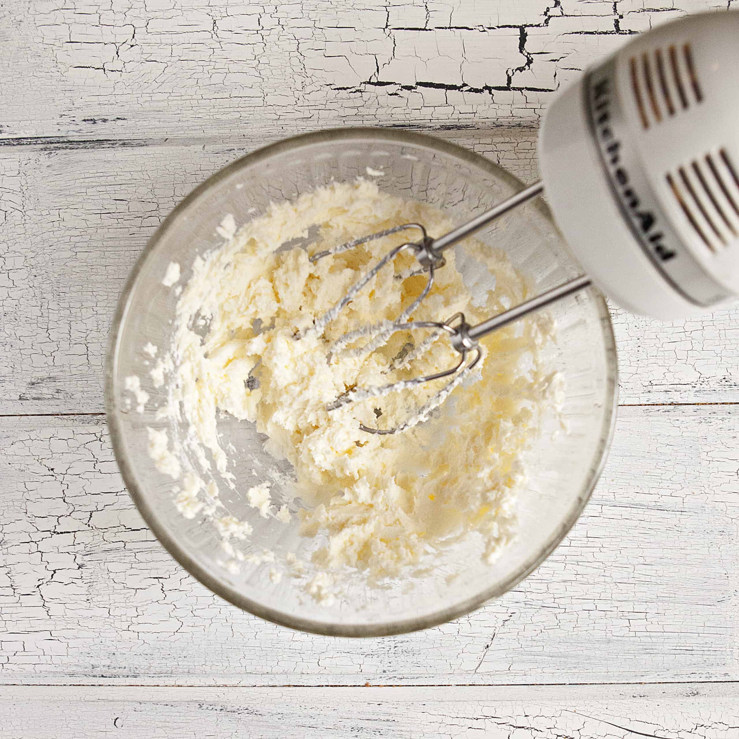 Butter and cream cheese being mixed by an electric mixer in a glass bowl on a white wood background.