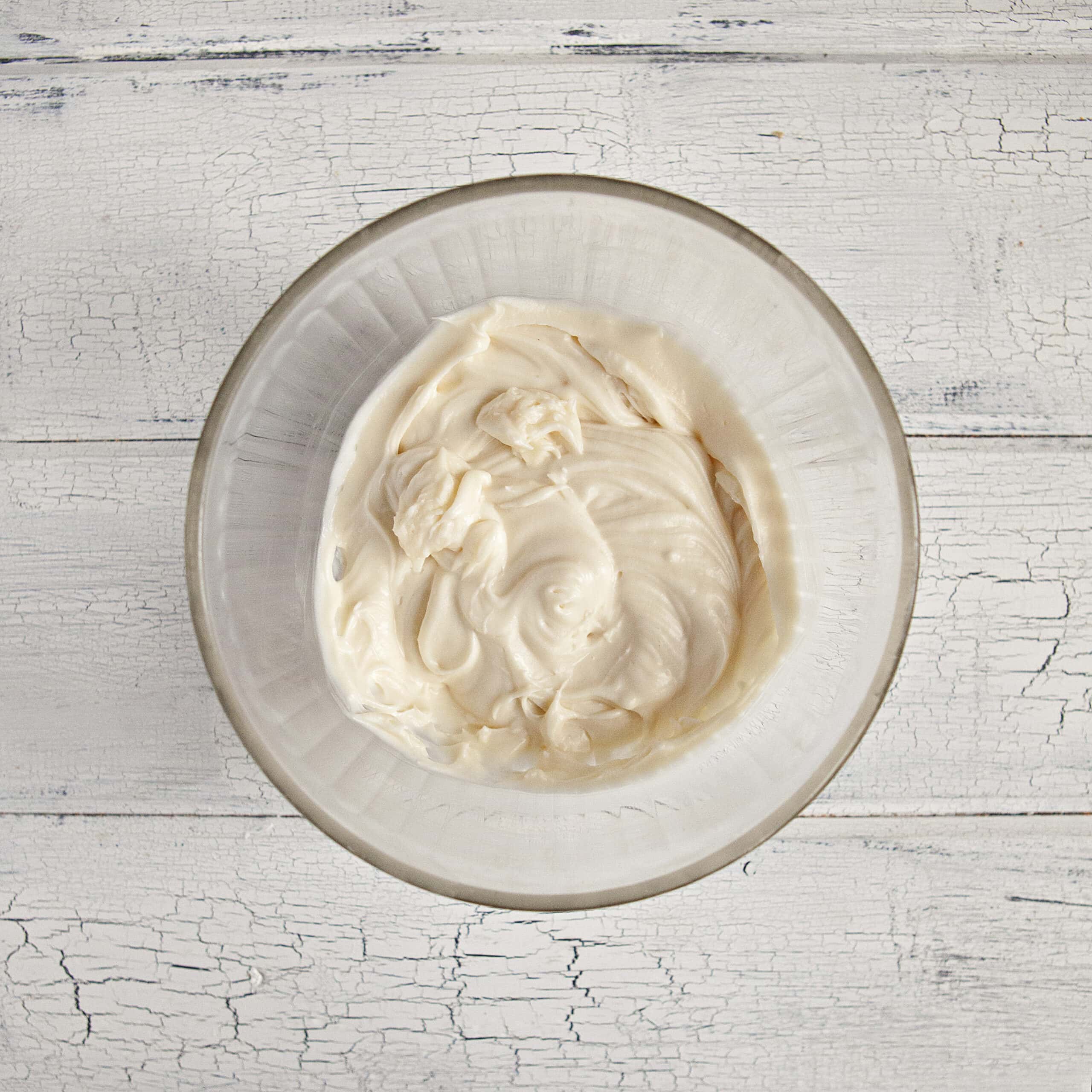 Gooey cream cheese frosting in a glass bowl on a white wood background.