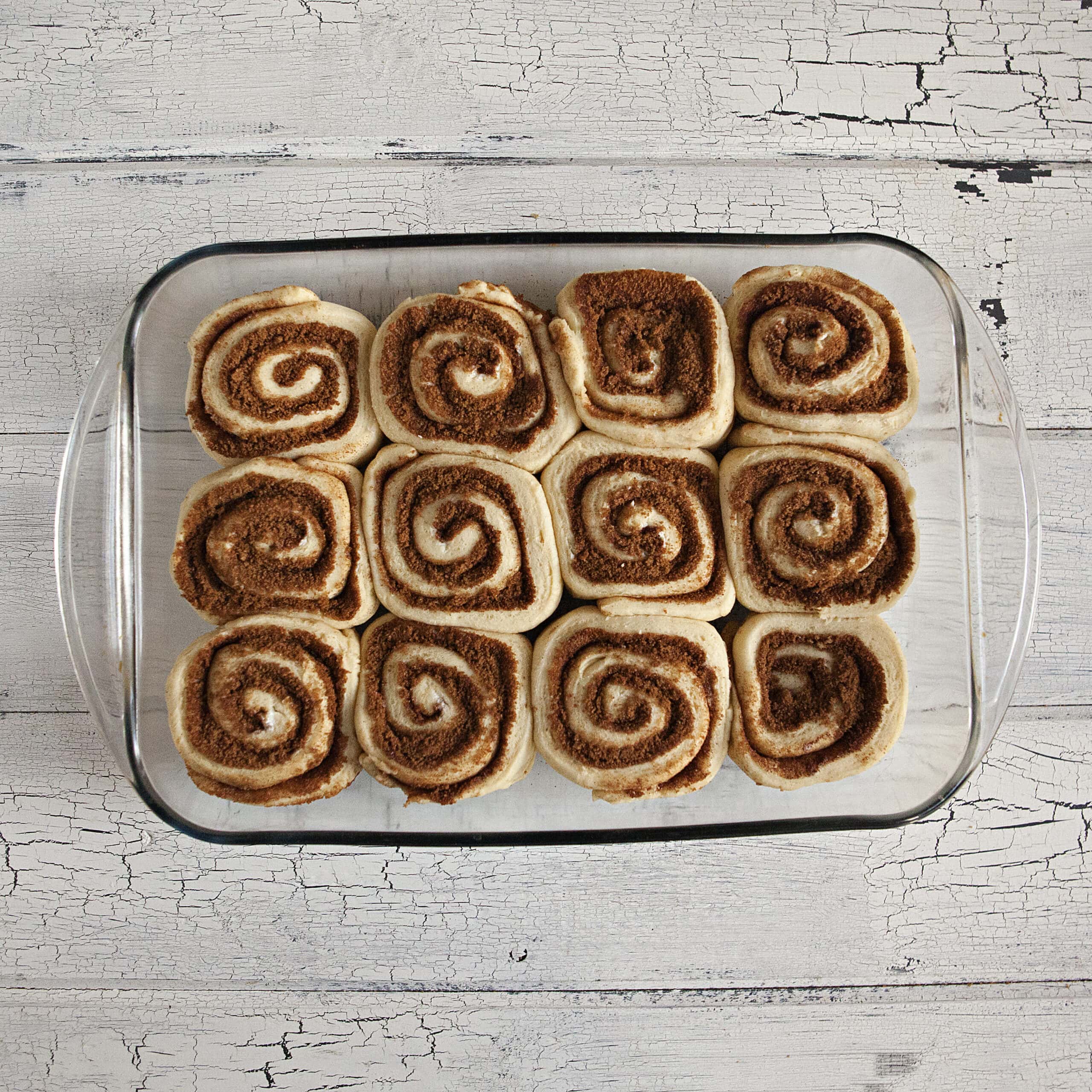 Risen uncooked soft and fluffy cinnamon rolls in a glass baking dish on a white wood background.