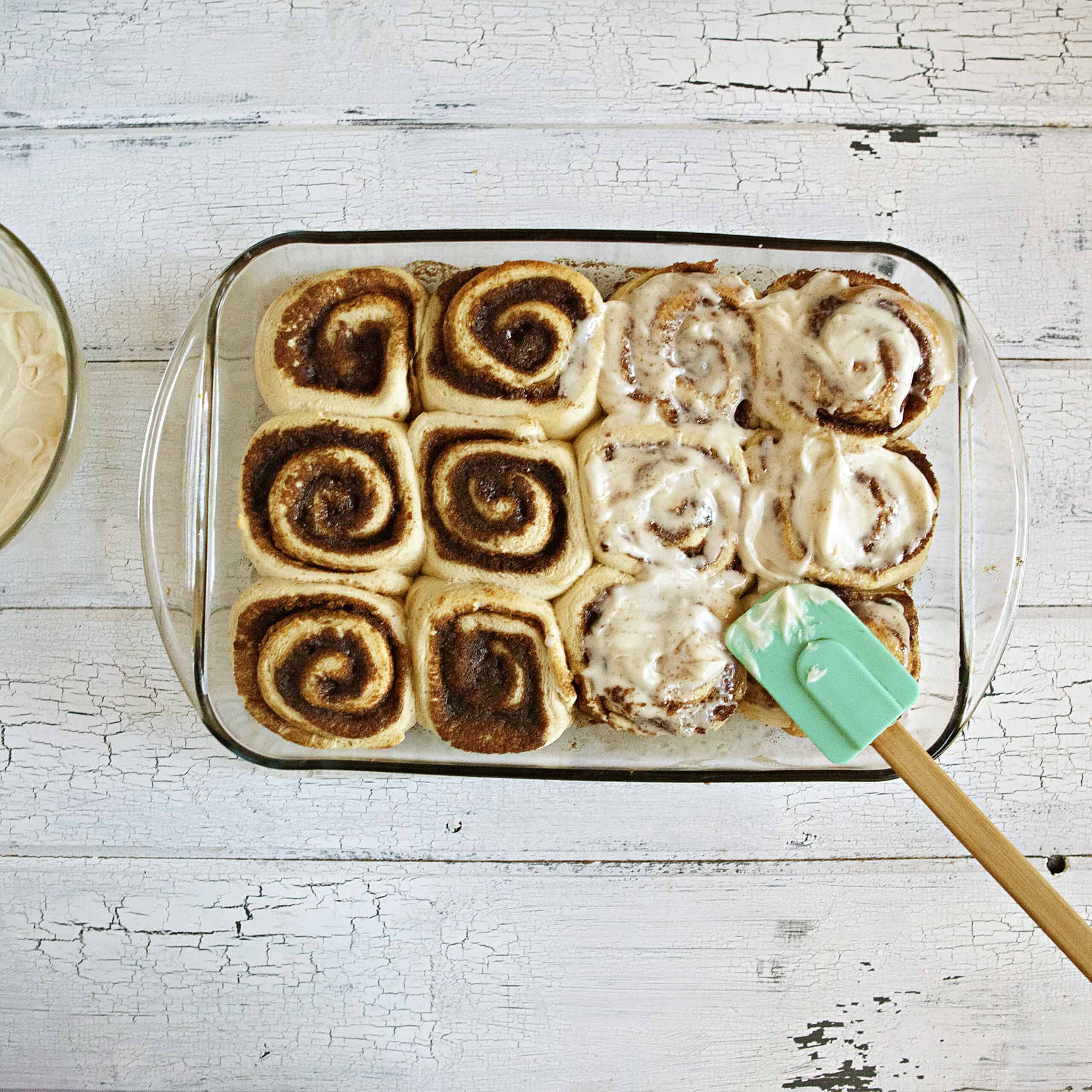 Baked soft and fluffy cinnamon rolls being frosted with cream cheese icing with a blue spatula in a glass baking dish on a white wood background.