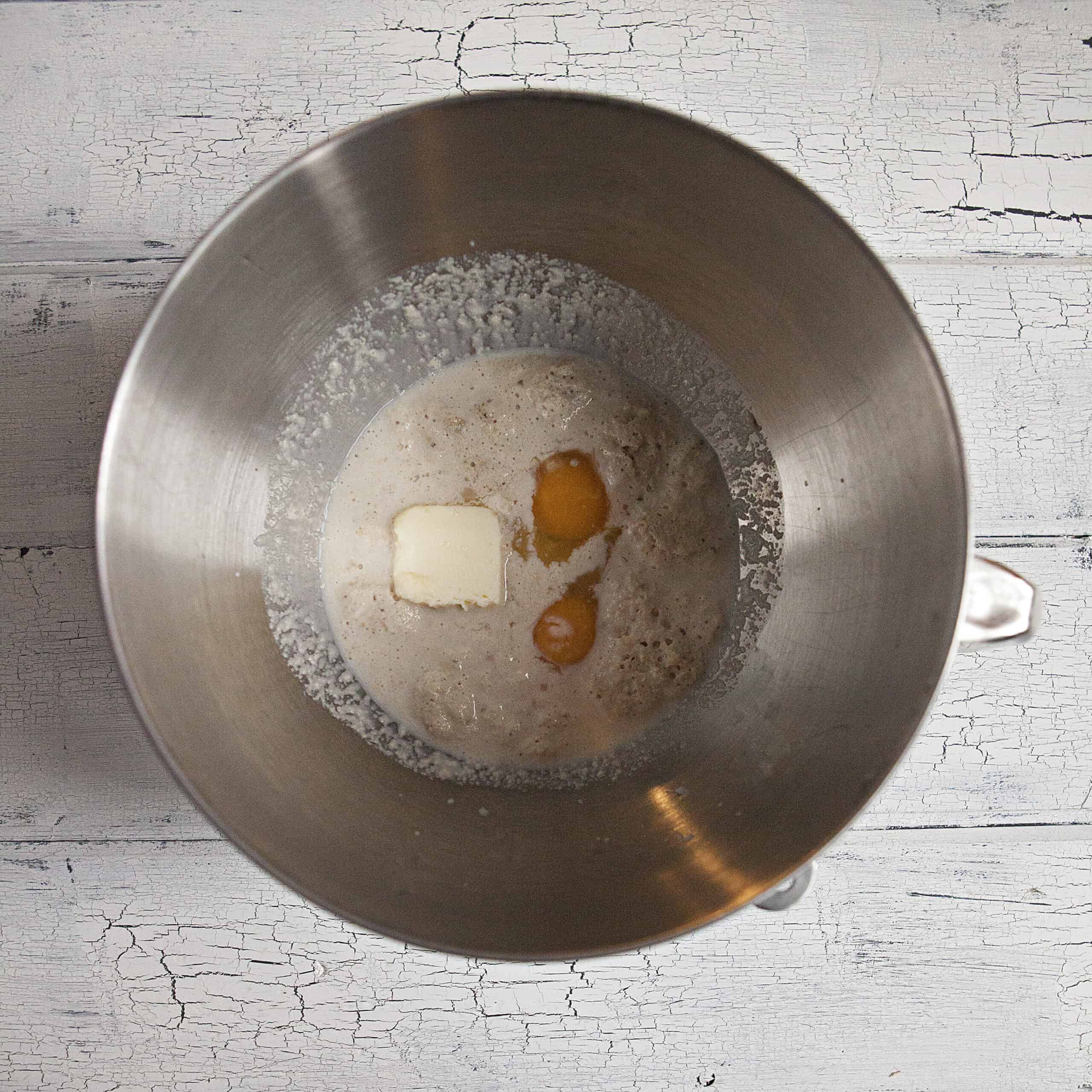 A stand mixer metal bowl containing milk, yeast, salt, butter, and eggs on a white wood background.