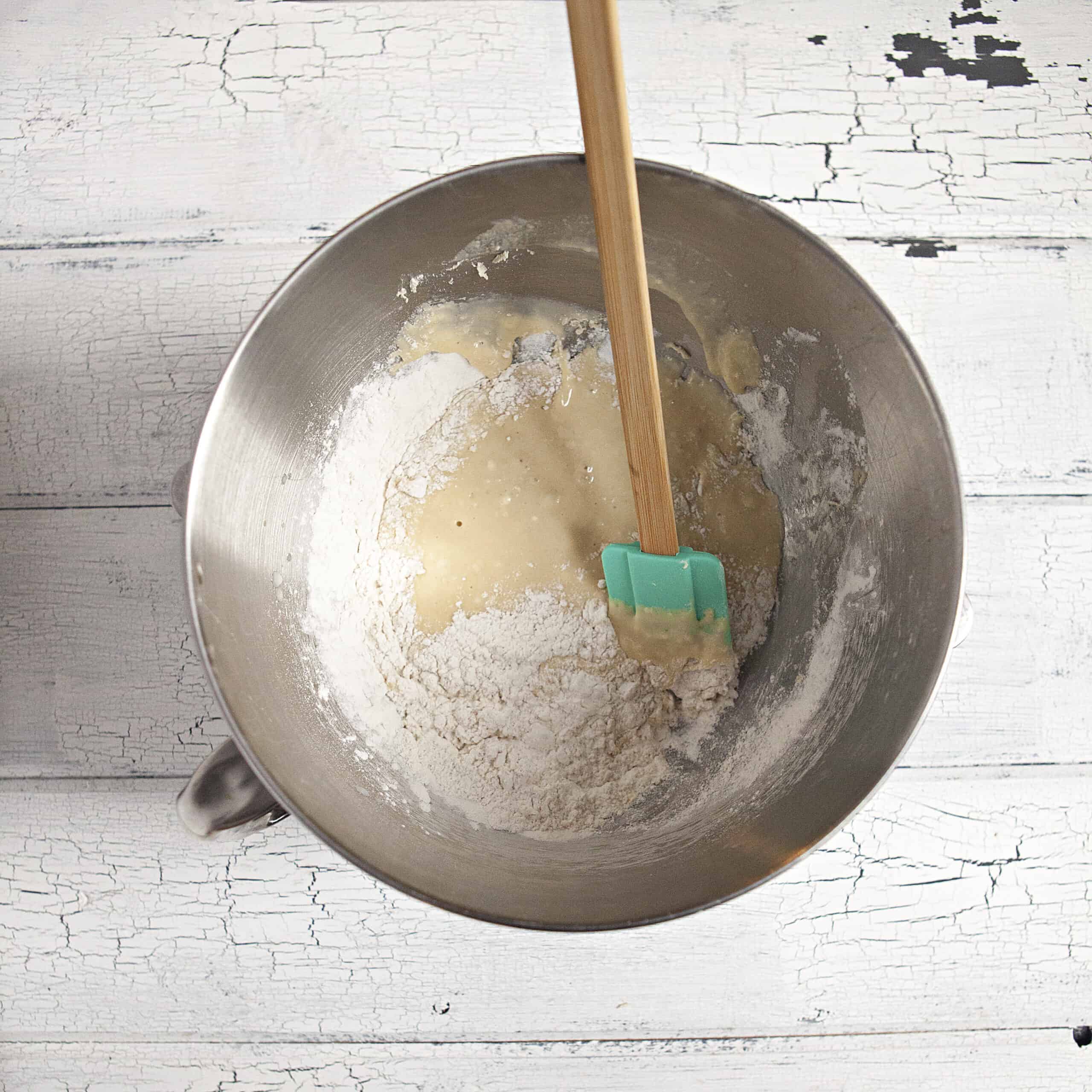 Ingredients for soft and fluffy cinnamon rolls in the metal bowl of a stand mixer with a blue spatula on a white wood background.