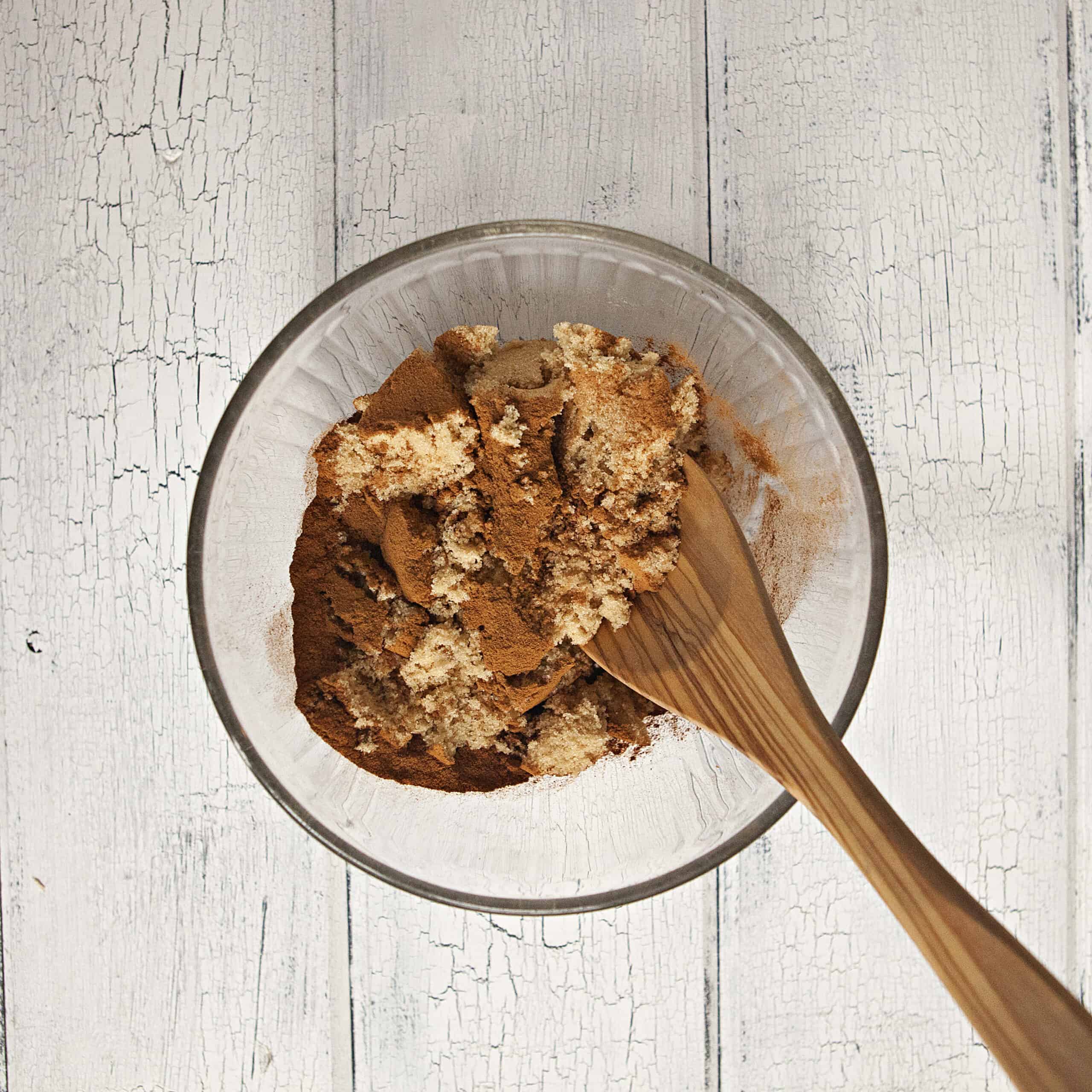 Brown sugar and cinnamon being mixed together with a wooden spoon in a glass bowl on a white wood background.