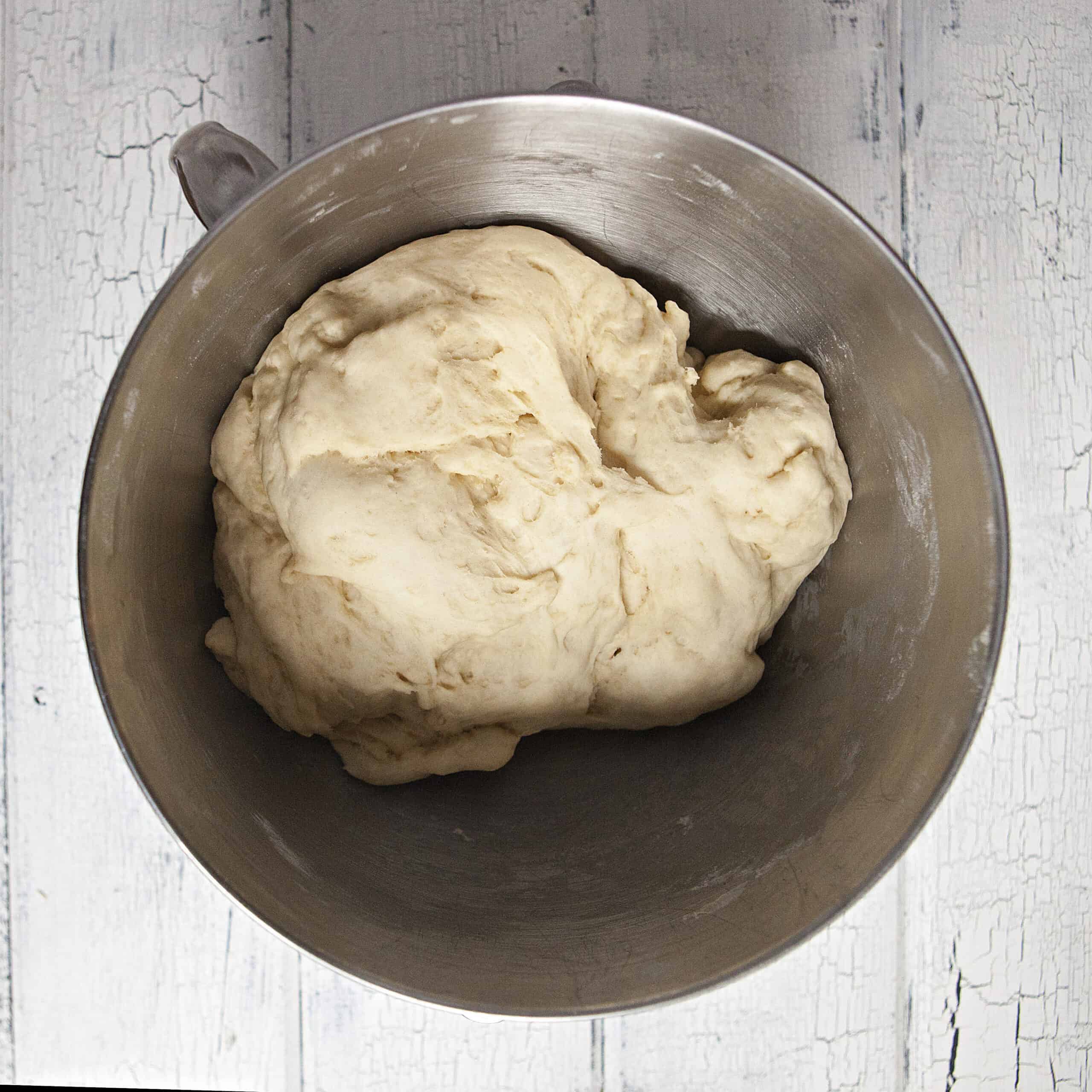 Risen dough for soft and pillow cinnamon rolls in the metal bowl of a stand mixer on a white wood background.