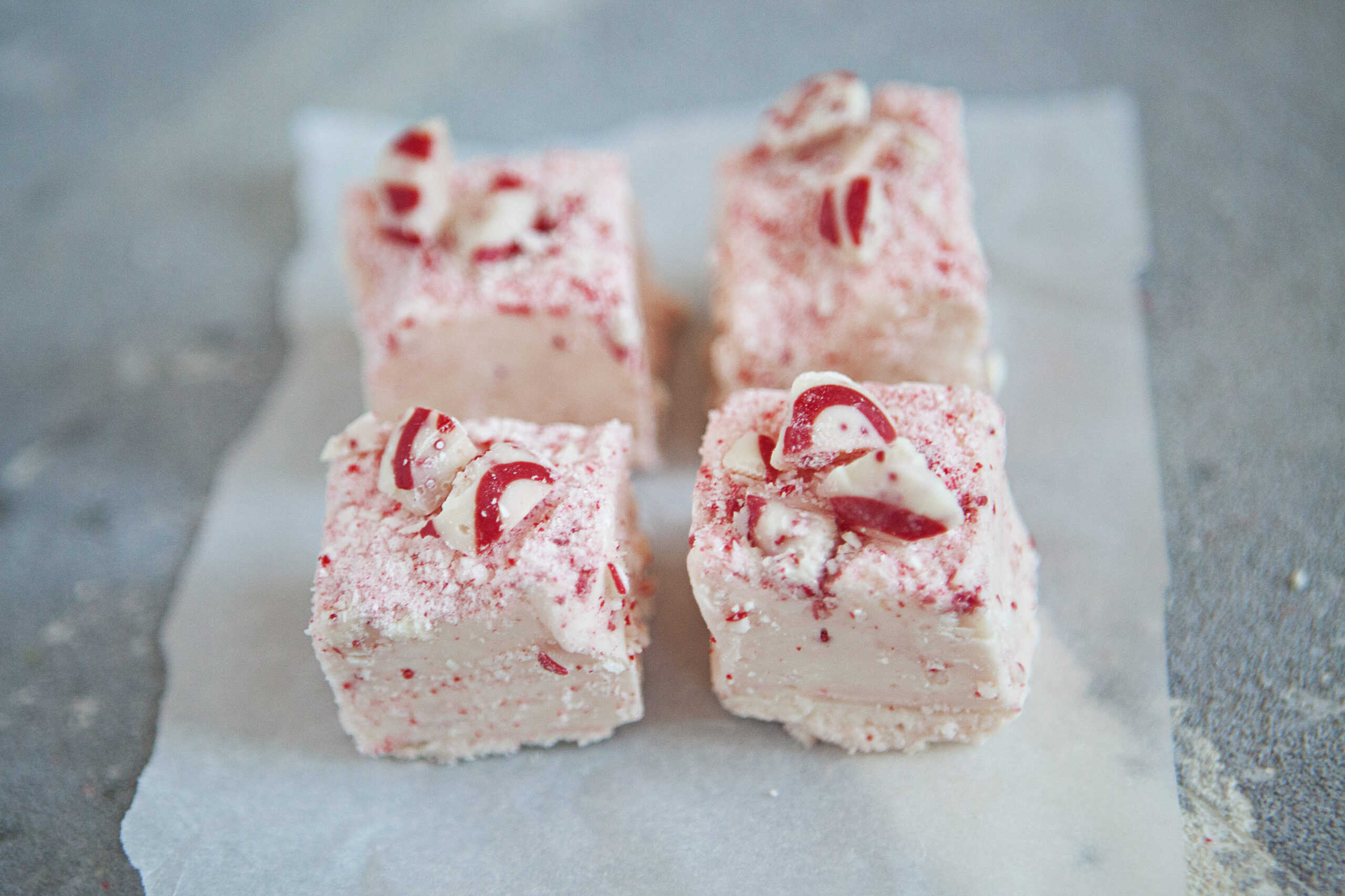 Four square pieces of red and white chocolate candy cane crunch fudge on a piece of parchment paper on a gray background.