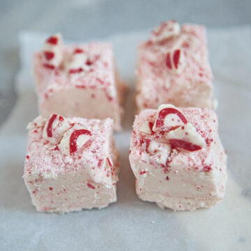 Four squares of red and white chocolate candy cane fudge on a square of parchment paper on a gray background.