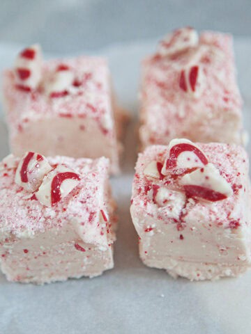 Four squares of red and white chocolate candy cane fudge on a square of parchment paper on a gray background.