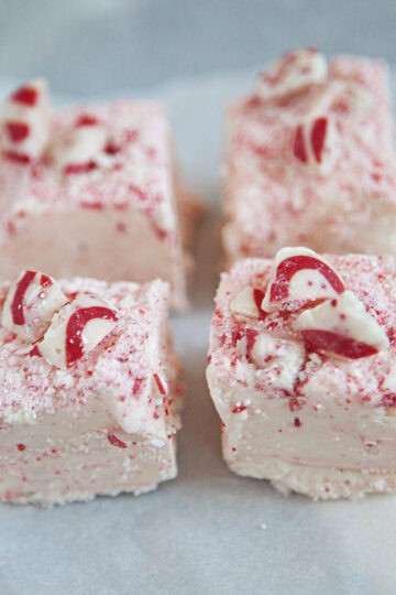 Four squares of red and white chocolate candy cane fudge on a square of parchment paper on a gray background.