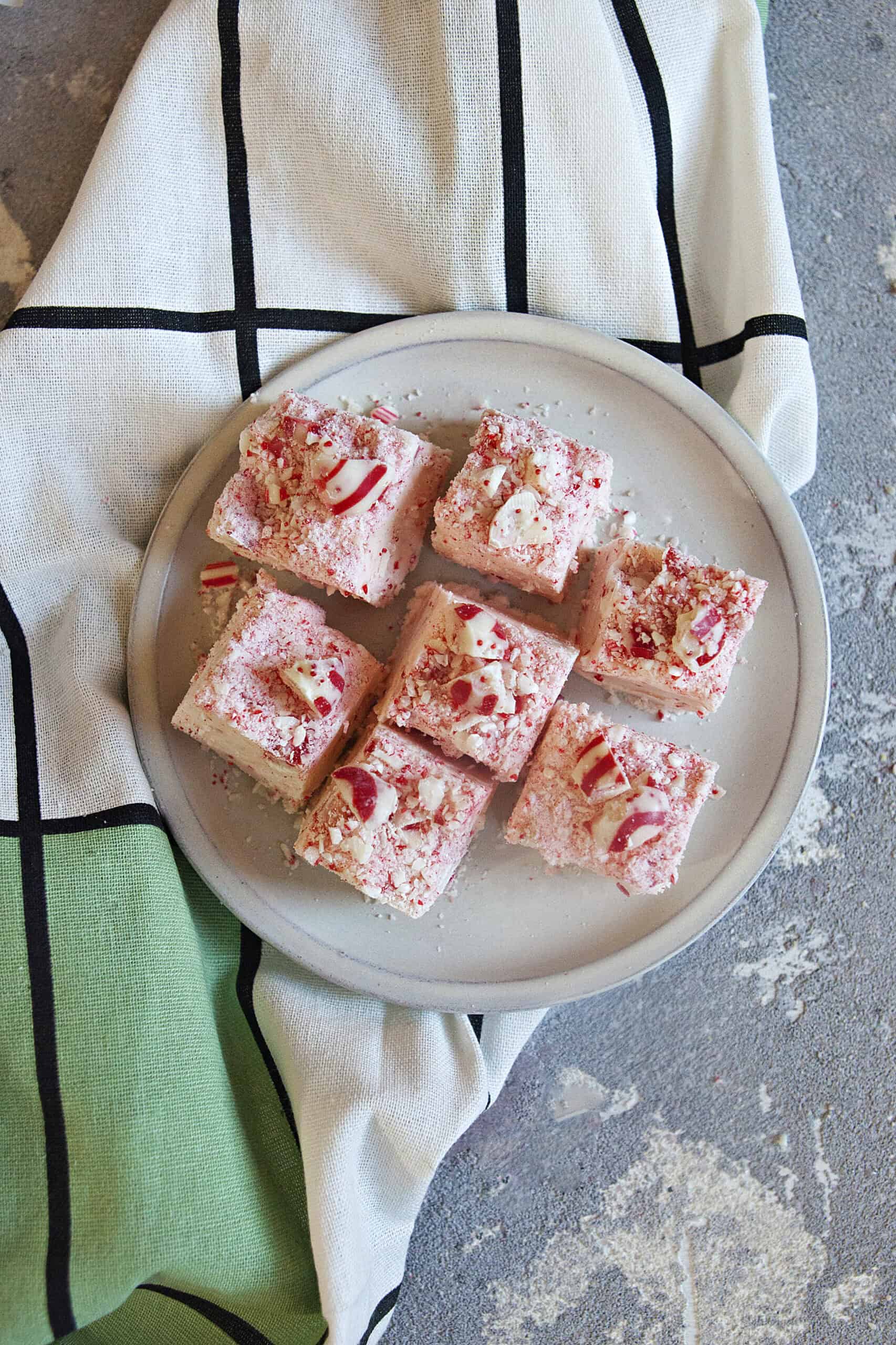 Seven pieces of pink, red, and white chocolate candy cane crunch fudge on an off-white plate alongside a green, white, and black checked kitchen towel on a gray background.