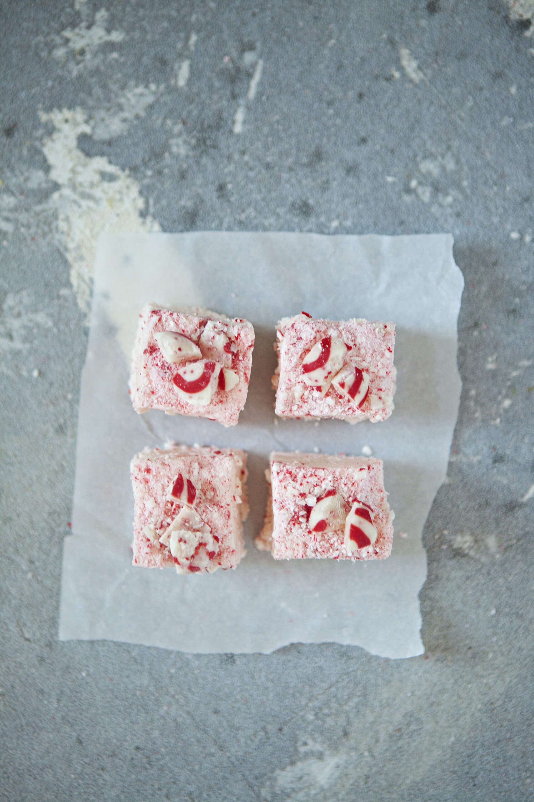 Four square pieces of pink, red, and white chocolate candy cane crunch fudge on a square of parchment paper on a gray background.