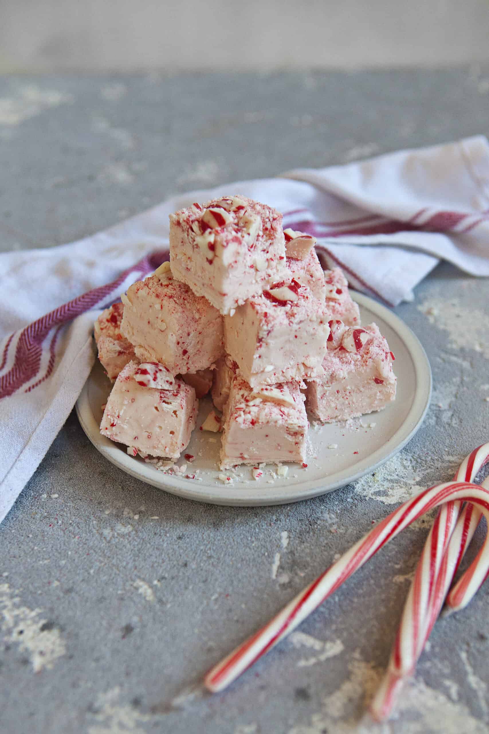 A pyramid of red, pink, and white chocolate candy cane crunch fudge on an off-white plate alongside a white and red kitchen towel and 3 whole candy canes on a gray background.