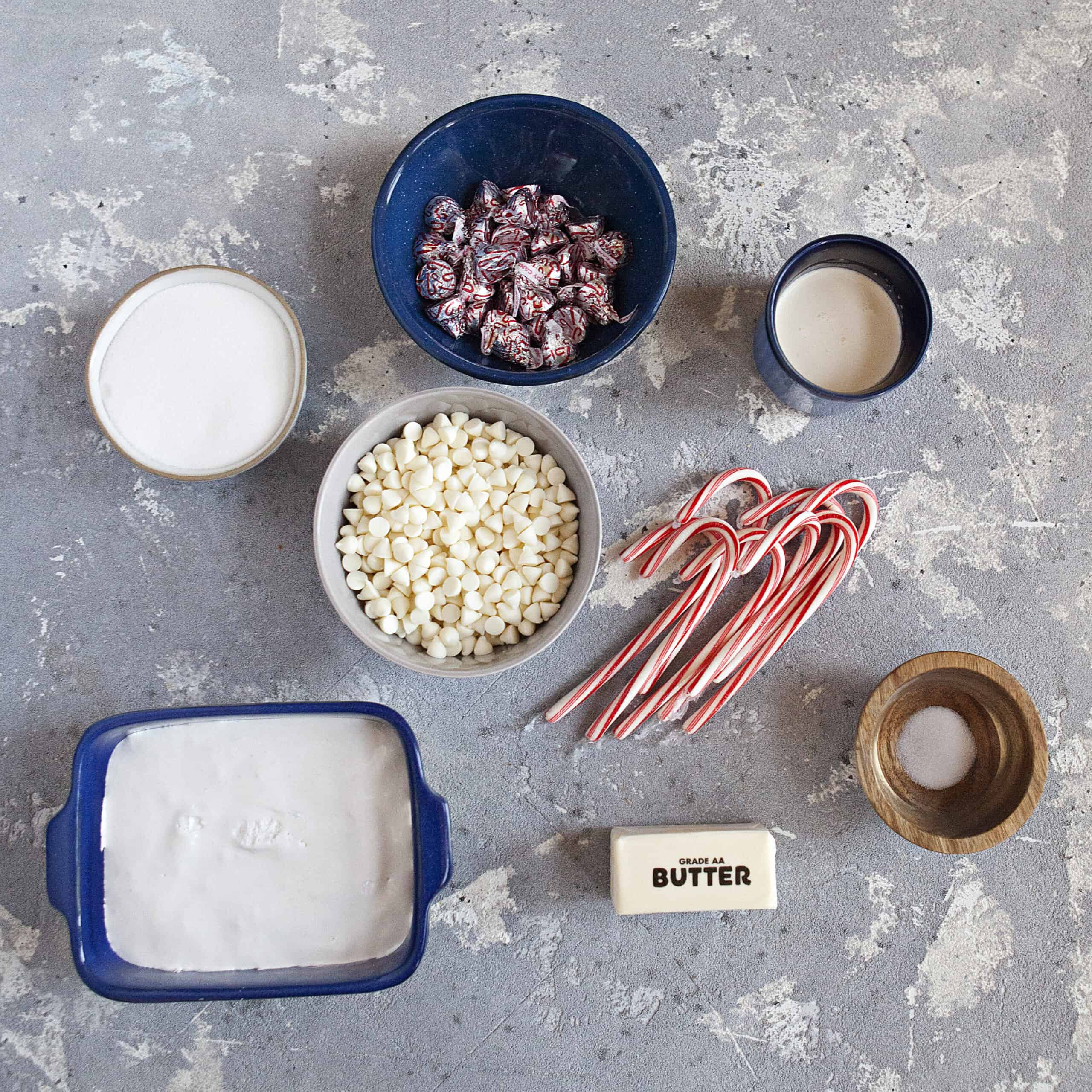 Ingredients for white chocolate candy cane crunch fudge:  Candy cane Hershey kisses, butter, sugar, heavy cream, white chocolate chips, marshmallow fluff, candy canes, and salt, on a gray backdrop.