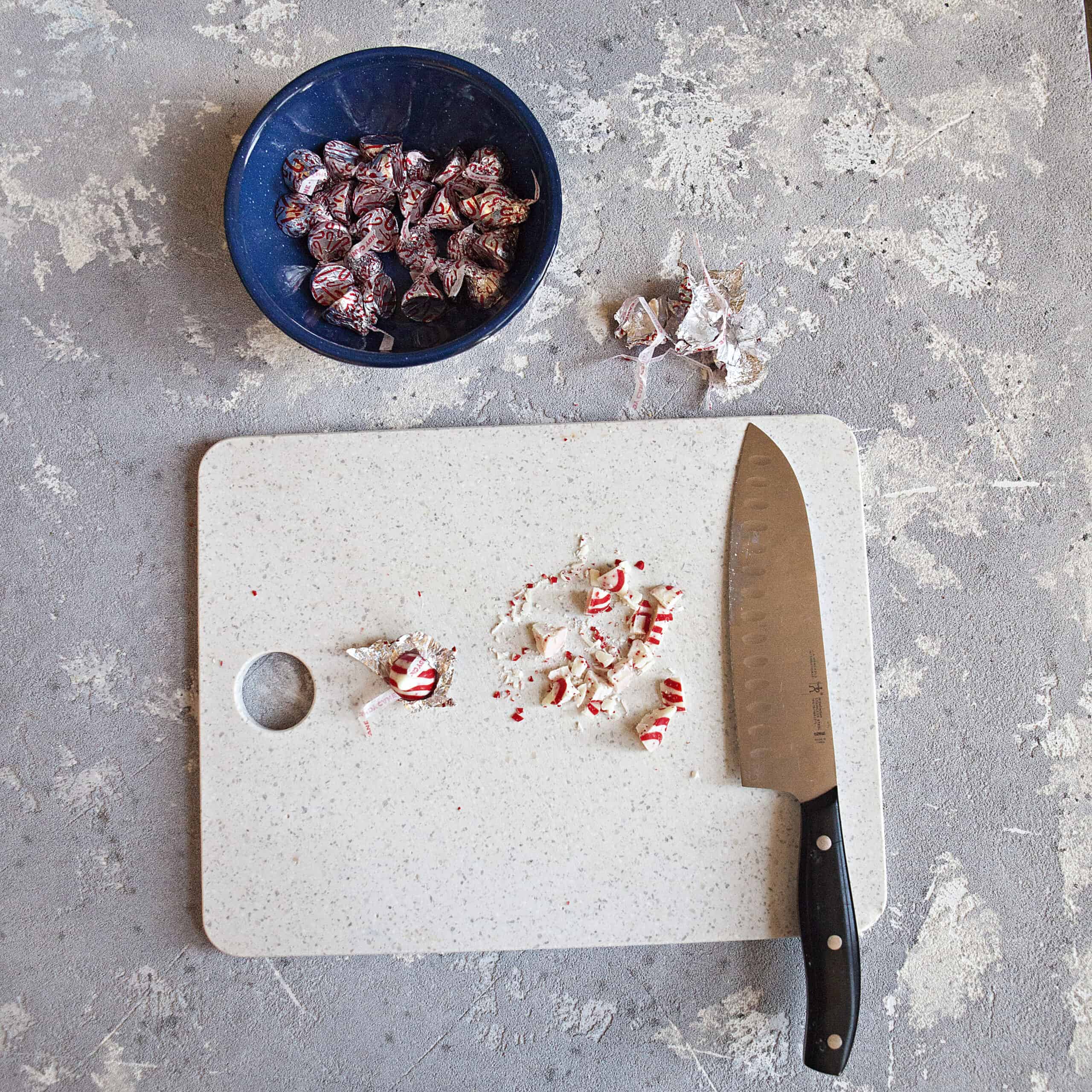 A bowl full of candy cane Hershey kisses next to a cutting board with a knife and chopped pieces of candy cane kisses and wrappers on a gray backdrop.