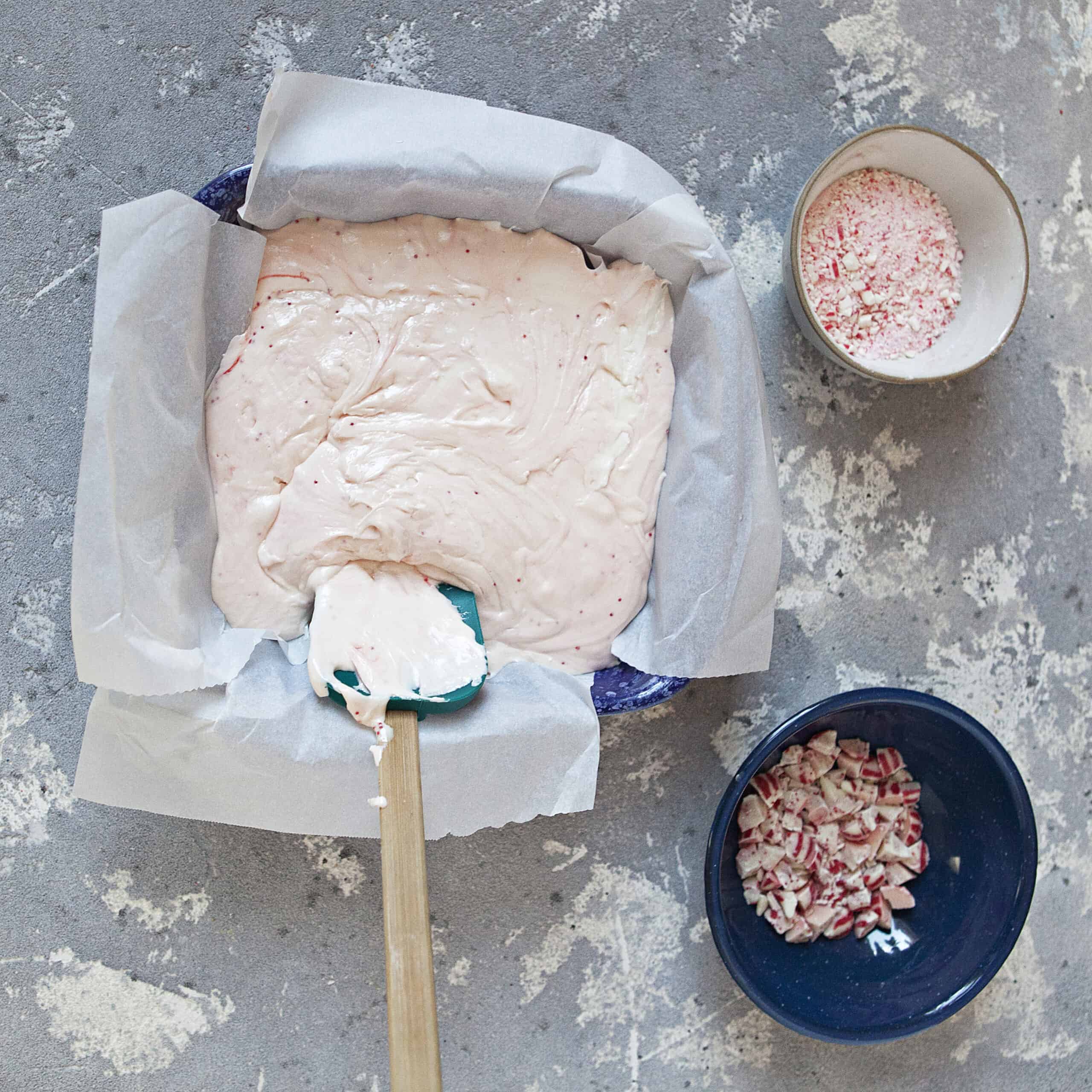 A 9x9-inch baking dish lined with parchment paper containing a light pink creamy mixture and a turquoise spatula, alongside 2 smaller bowls containing candy cane powder and chunks and chopped red and white candy cane Hershey kisses, on a gray background.