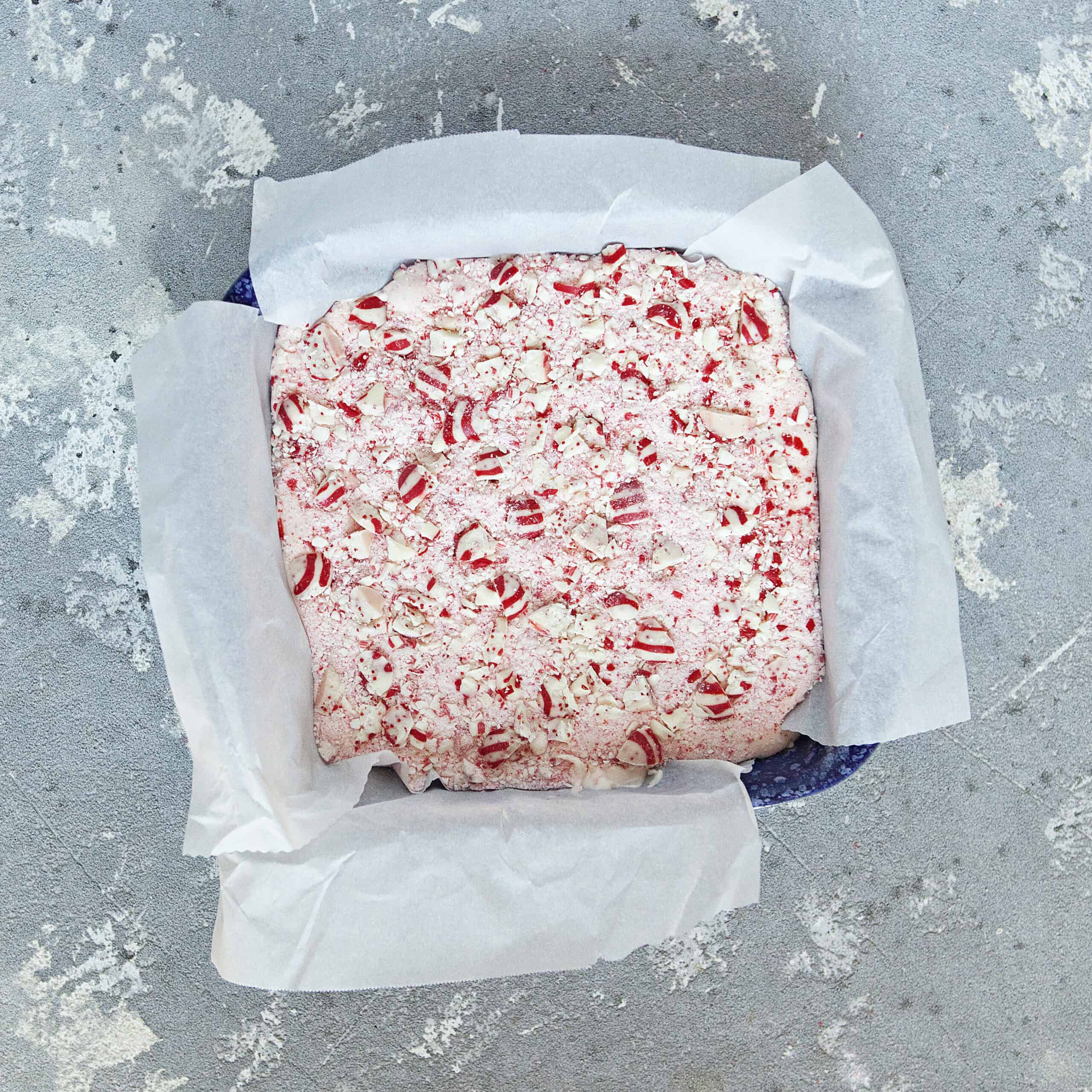 A 9x9-inch baking dish lined with parchment paper containing a light pink creamy mixture topped with red and white striped chopped candy cane Hershey kisses and powdered candy canes on a gray background.