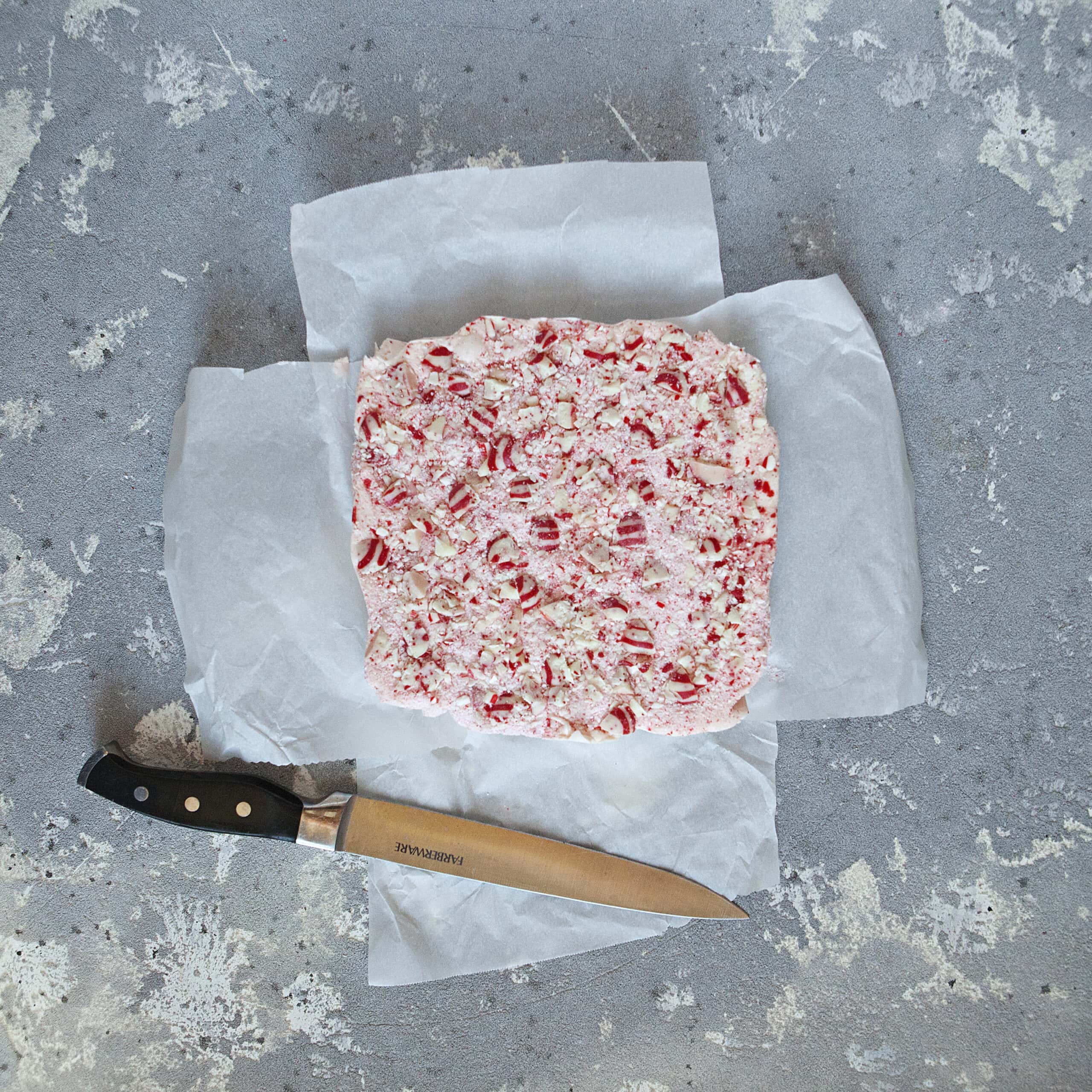 A light pink block of white chocolate candy cane crunch fudge with red and white striped candies on 2 sheets of parchment paper alongside a chef's knife on a gray background.