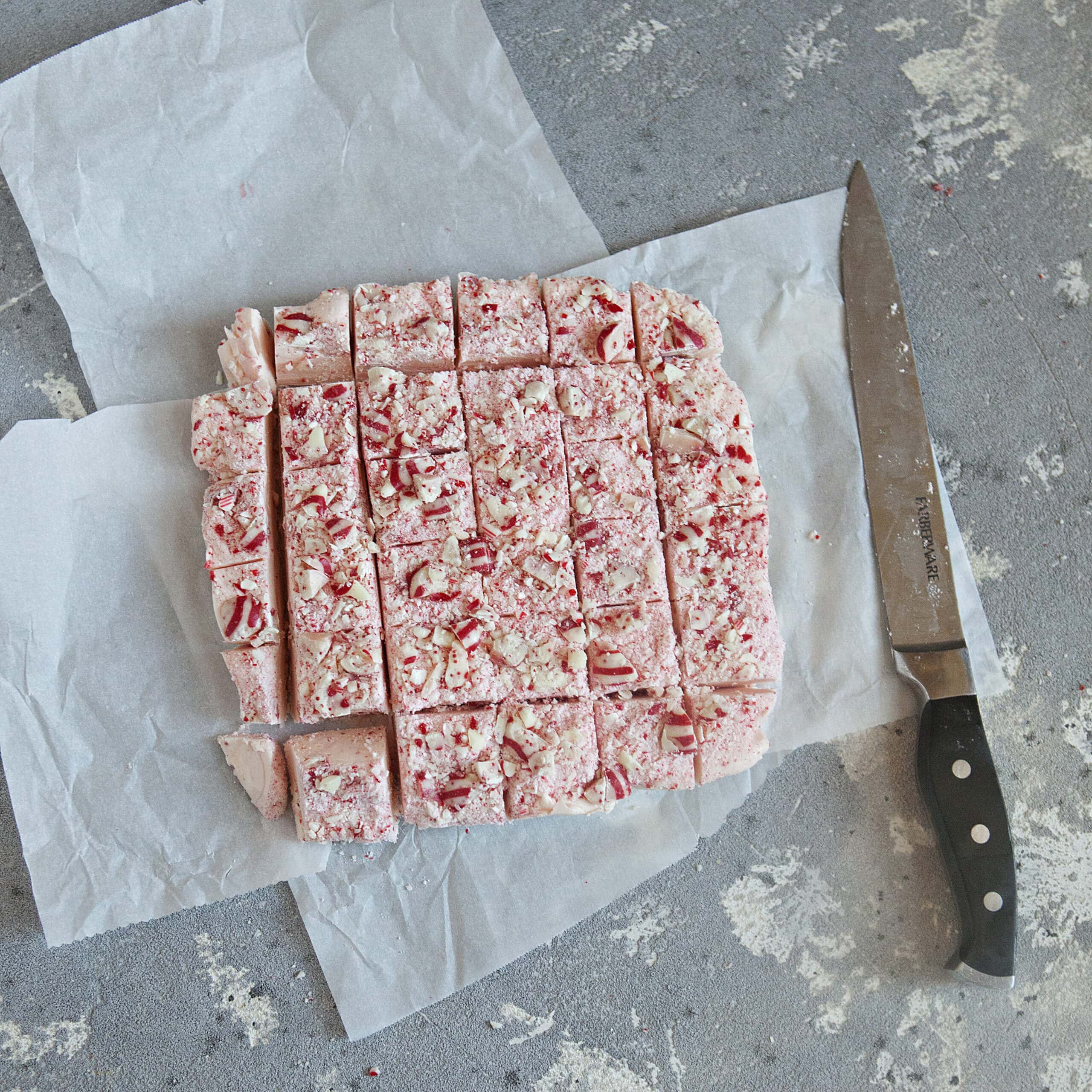 A light pink block of white chocolate candy cane crunch fudge topped with red and white striped candy cane Hershey kisses and powdered candy canes, chopped into 36 equal pieces, alongside a chef's knife on a gray background.