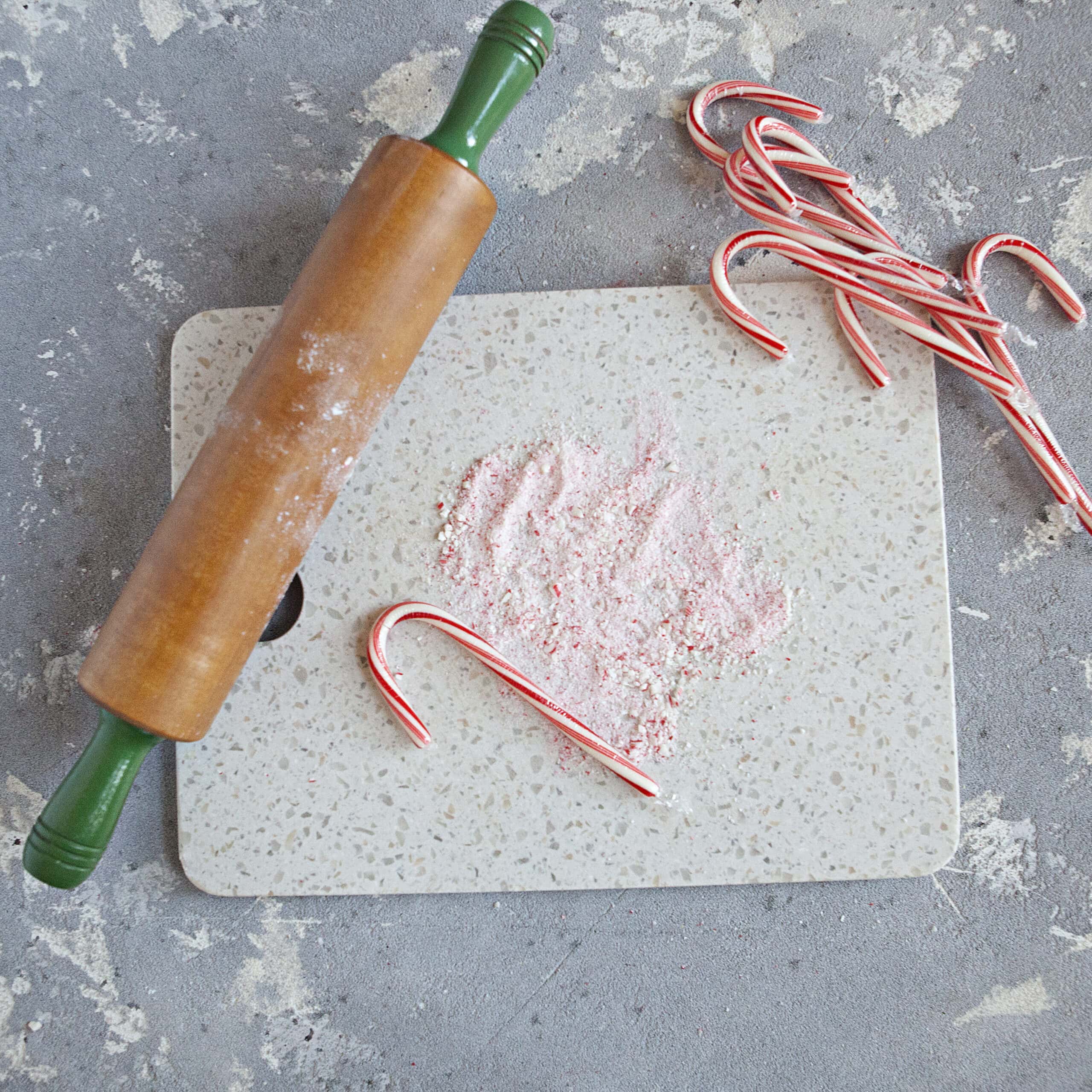 A red and white striped candy cane on a cutting board next to powdered candy cane, a rolling pin, and several intact candy canes on a gray background.