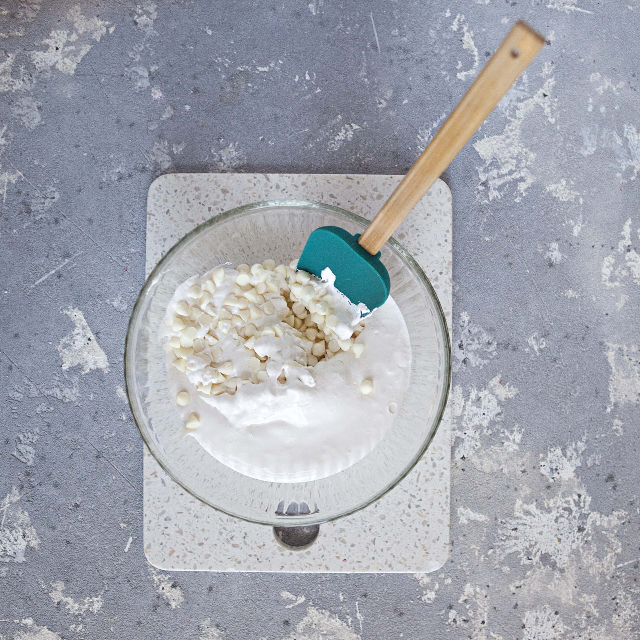 A glass bowl containing white chocolate chips, marshmallow fluff, and a turquoise spatula on a white cutting board and a gray background.