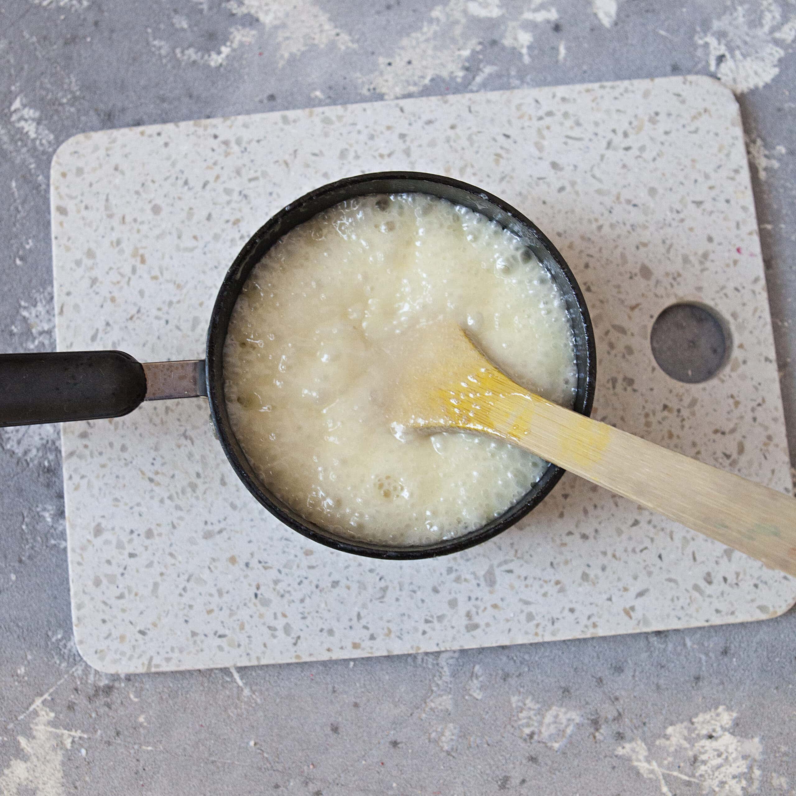 A saucepan containing a bubbly white mixture and a wooden spoon on a white cutting board and a gray background.