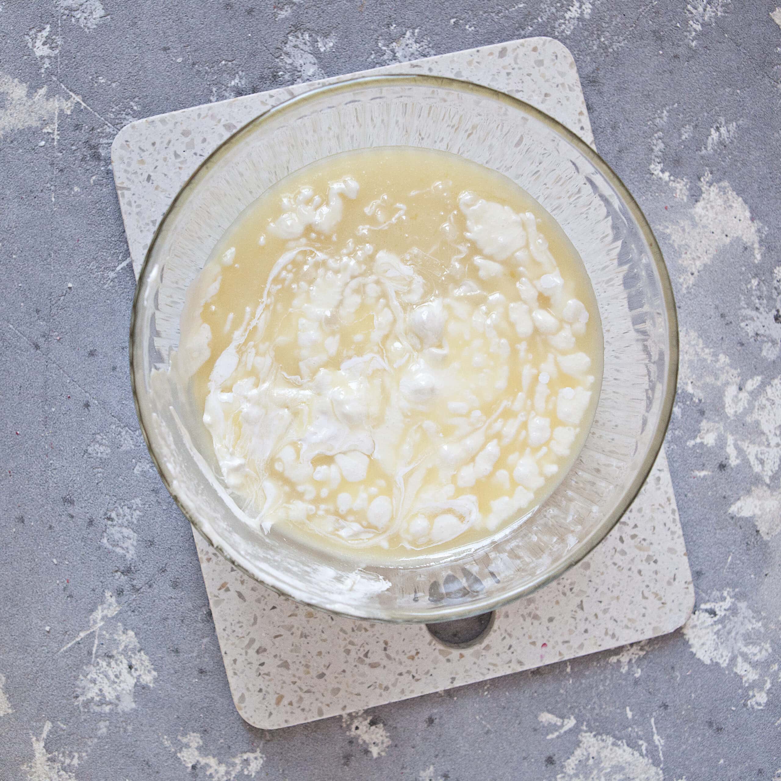 A glass bowl containing a tan and white mixture on a white cutting board and a gray background.