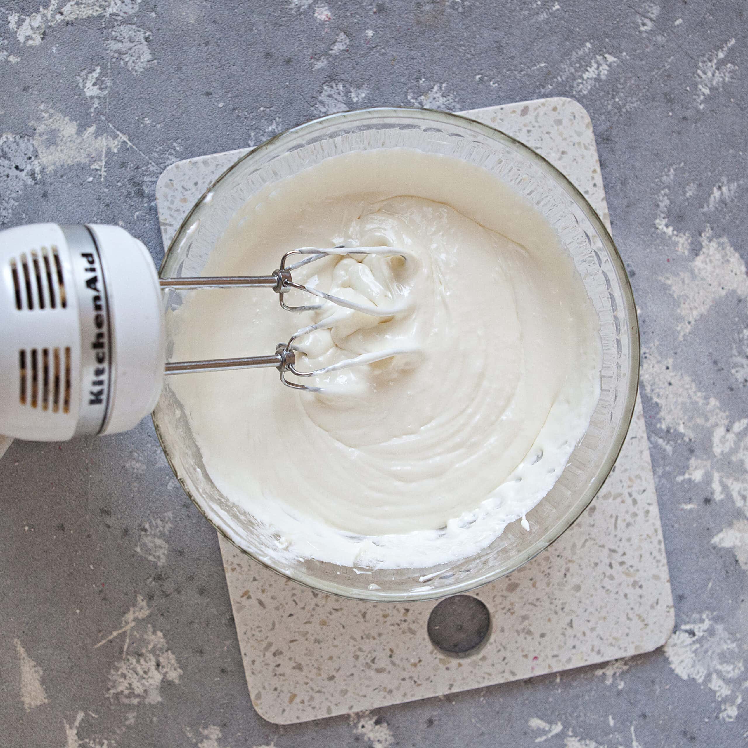 A glass bowl containing a creamy white mixture alongside an electric mixer with beaters mixing the contents of the bowl, on top of a white cutting board and a gray background.