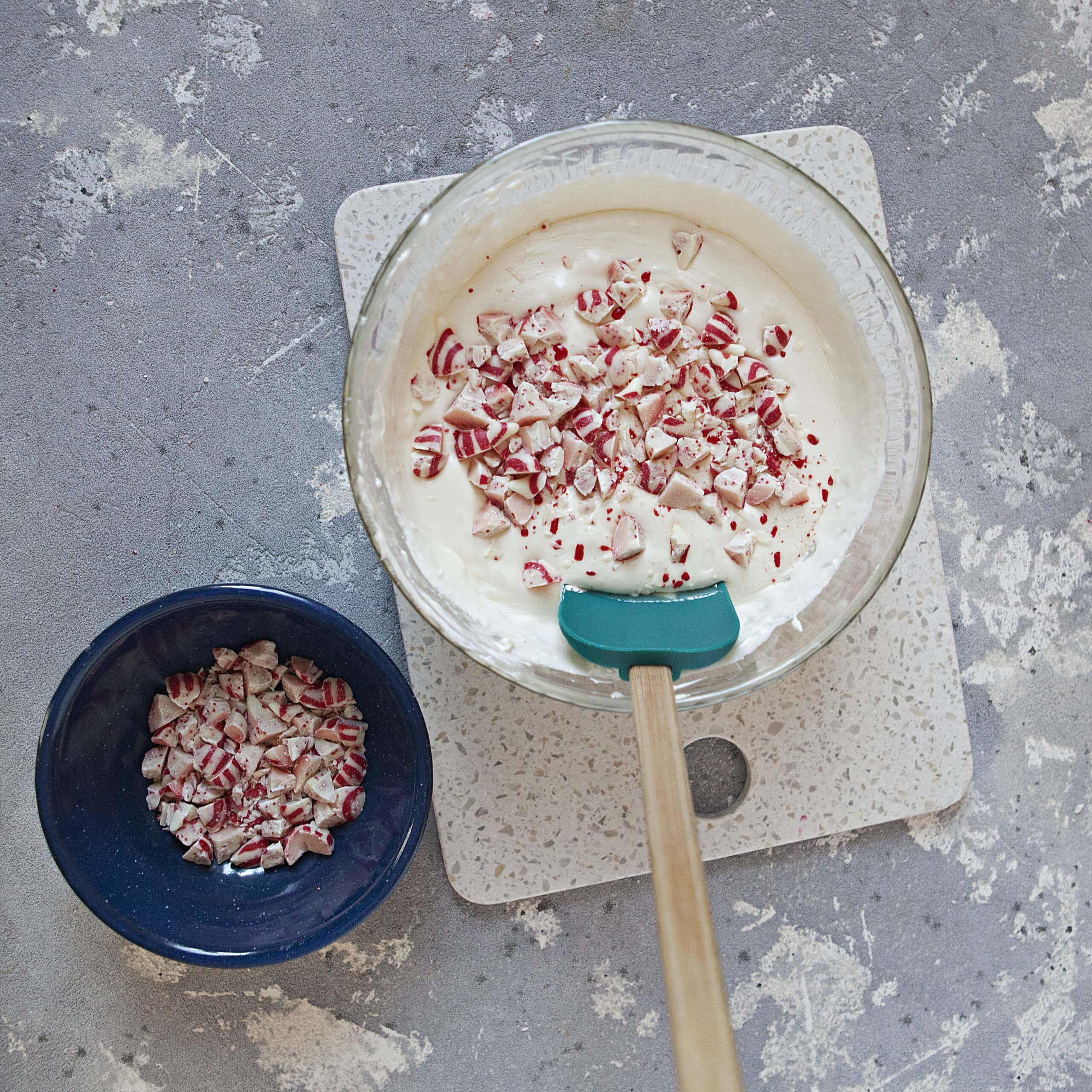 A glass bowl containing a creamy white mixture topped with chopped red and white striped candy cane Hershey kisses and a turquoise spatula, alongside a smaller blue bowl containing more chopped candy cane kisses, on a white cutting board and a gray background.