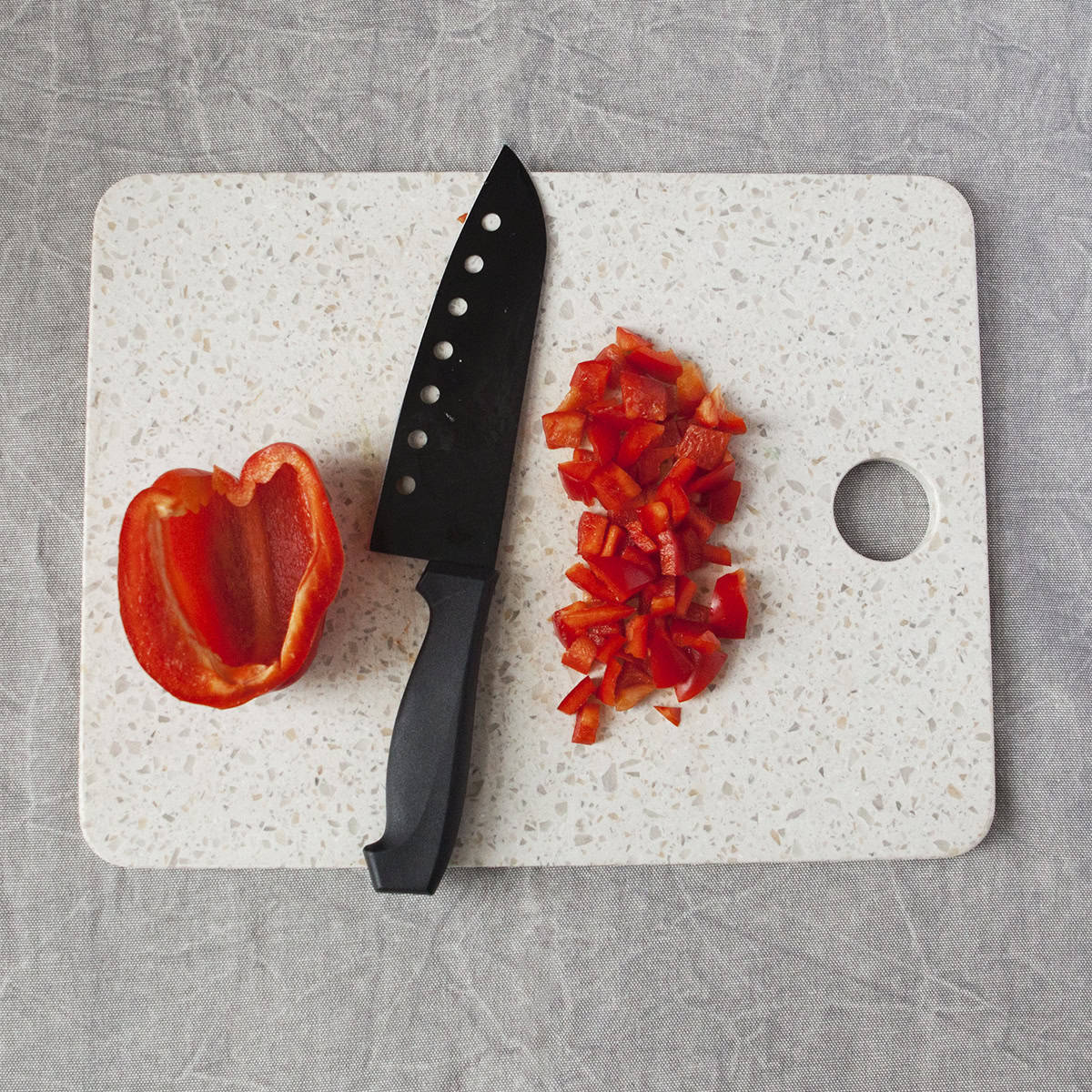 Half of a red bell pepper next to a black chef's knife and chopped red bell pepper on a white cutting board on a gray background.