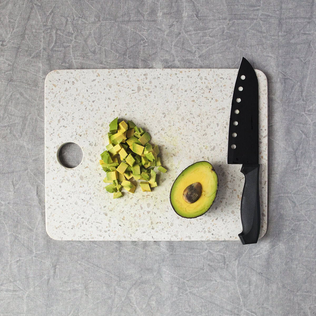 Half of an avocado next to chopped avocado with a black chef's knife on a white cutting board on a gray background.