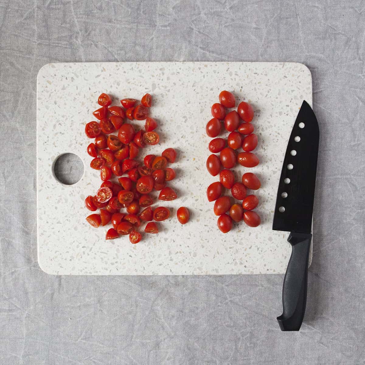 Whole cherry tomatoes and halved cherry tomatoes next to a black chef's knife on a white cutting board on a gray background.