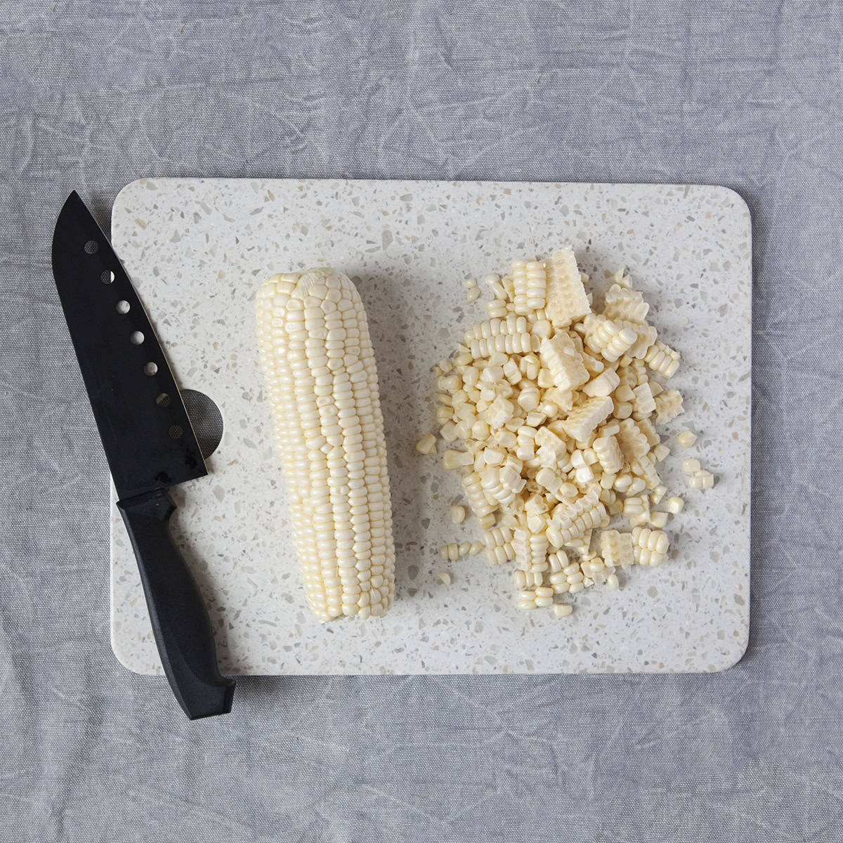 An ear of fresh corn next to a pile of corn kernels on a white cutting board with a black chef's knife on a gray background.