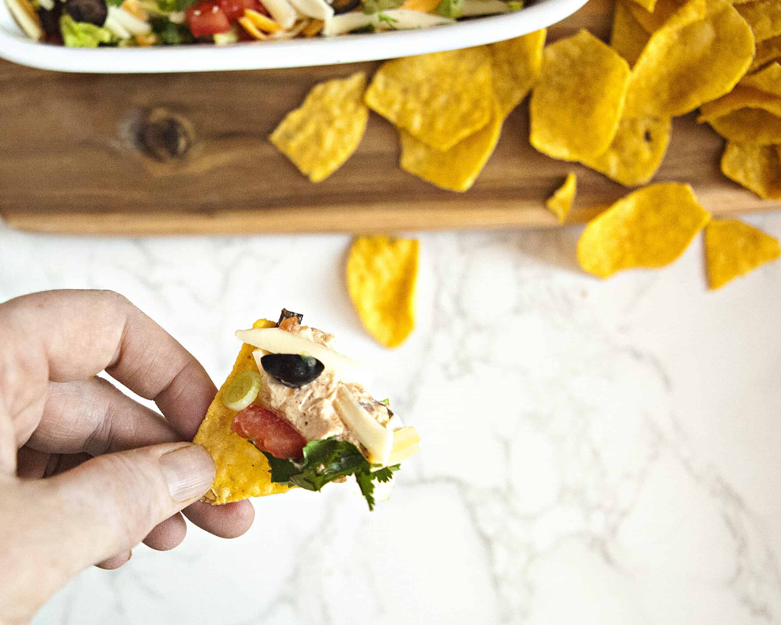 Cream cheese taco dip on a tortilla chip being held by a hand, alongside more dip in a baking dish and tortilla chips.