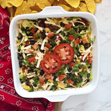 Quick cream cheese taco dip topped with shredded cheese, green onions, sliced black olives, diced tomatoes, cilantro, and 2 tomato slices in a square white baking dish next to a red bandanna-patterned kitchen towel and tortilla chips.