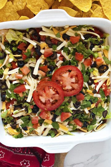 Quick cream cheese taco dip topped with shredded cheese, green onions, sliced black olives, diced tomatoes, cilantro, and 2 tomato slices in a square white baking dish next to a red bandanna-patterned kitchen towel and tortilla chips.