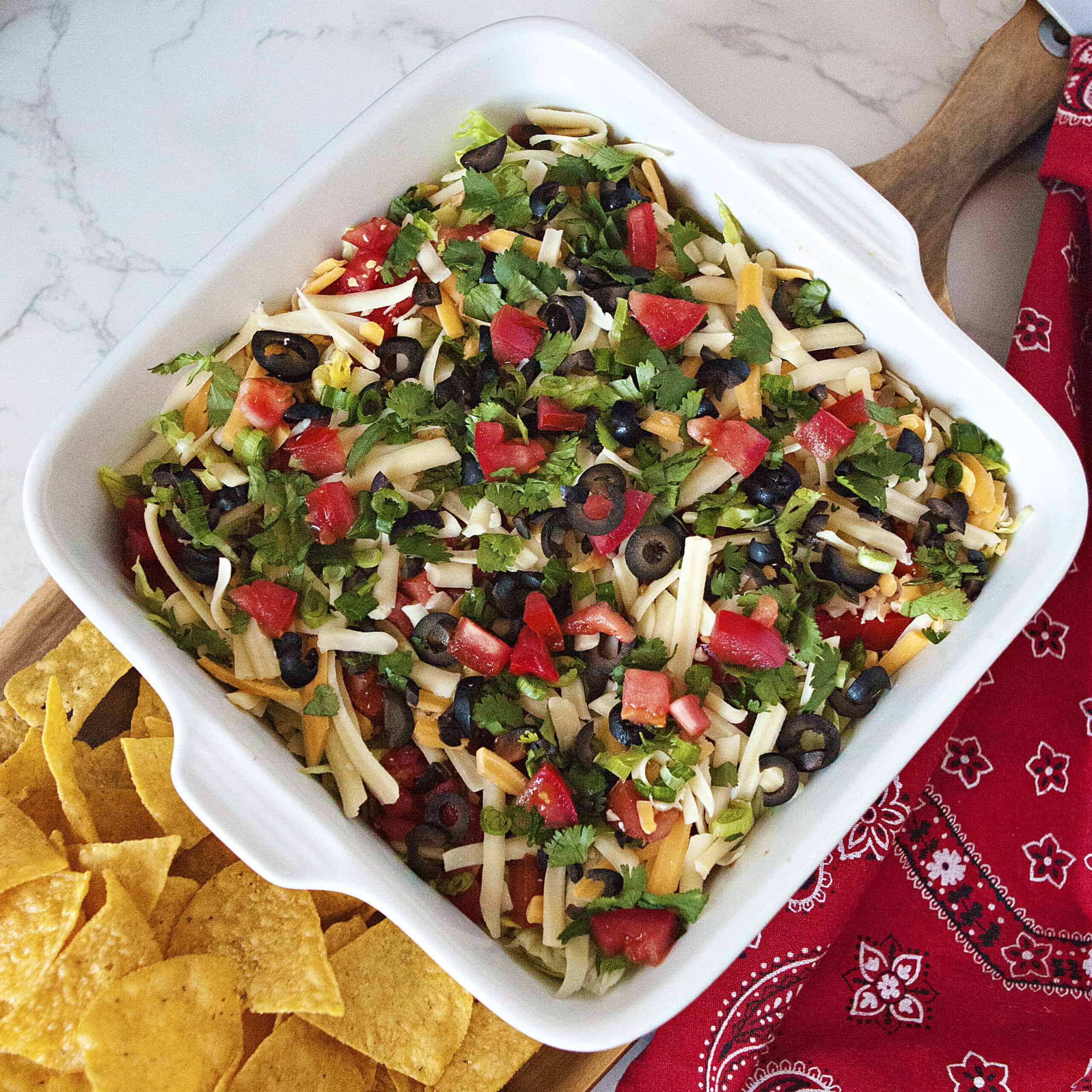 Quick cream cheese taco dip topped with lettuce, tomatoes, olives, cilantro, cheese, and green onions in a baking dish alongside a red kitchen towel and tortilla chips.