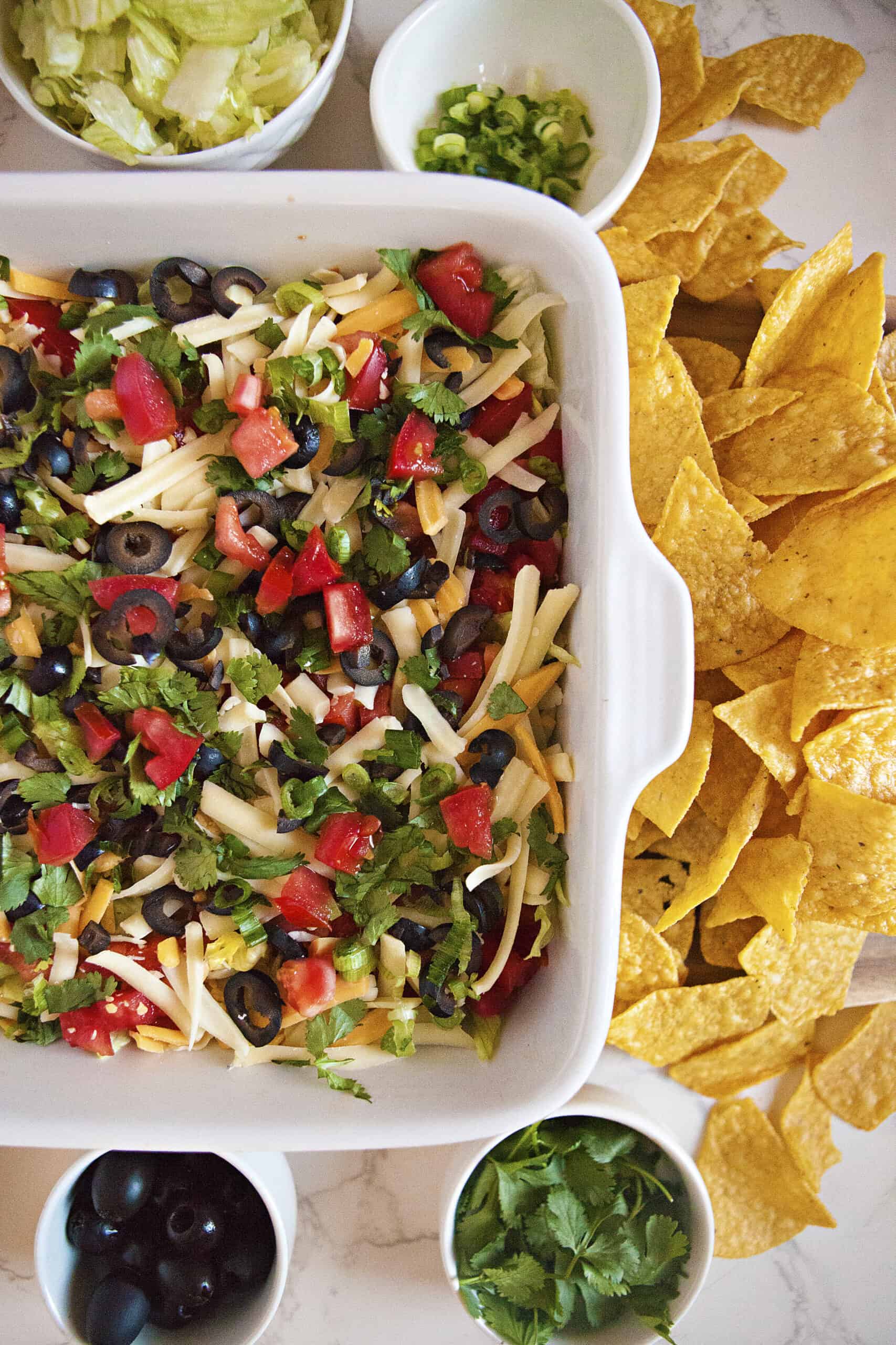 Cream cheese taco dip in a square baking dish alongside small dishes of black olives, cilantro, chopped lettuce, and chopped green onions as well as nacho chips.