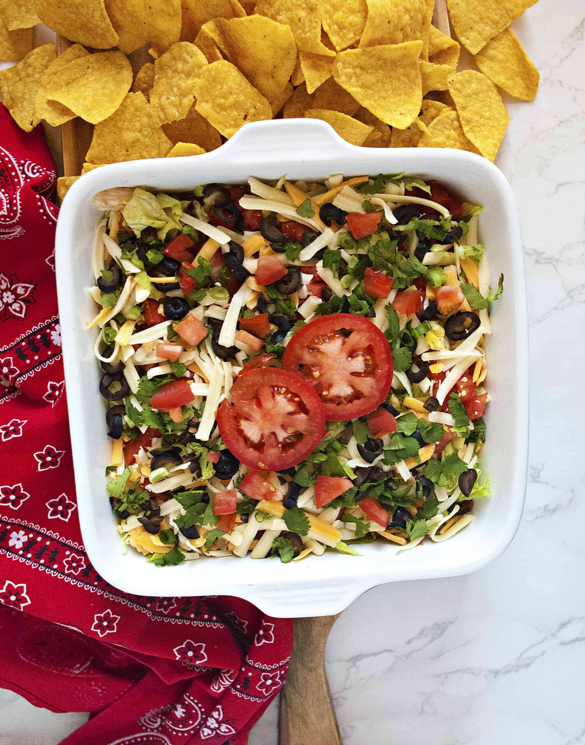 Quick cream cheese taco dip topped with shredded cheese, green onions, sliced black olives, diced tomatoes, cilantro, and 2 tomato slices in a square white baking dish next to a red bandanna-patterned kitchen towel and tortilla chips.