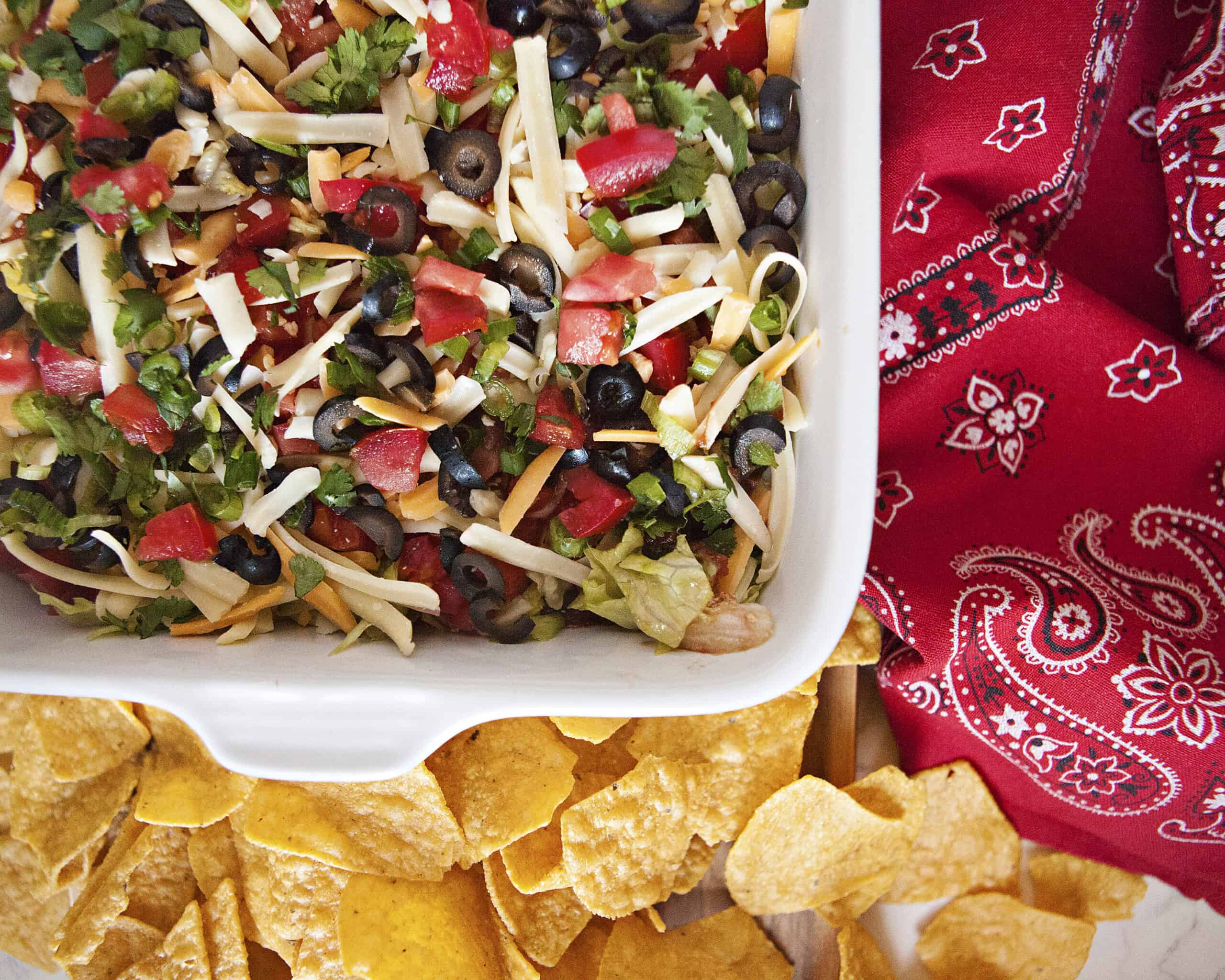 Quick cream cheese taco dip topped with lettuce, tomatoes, olives, cilantro, cheese, and green onions in a baking dish alongside a red kitchen towel and tortilla chips.