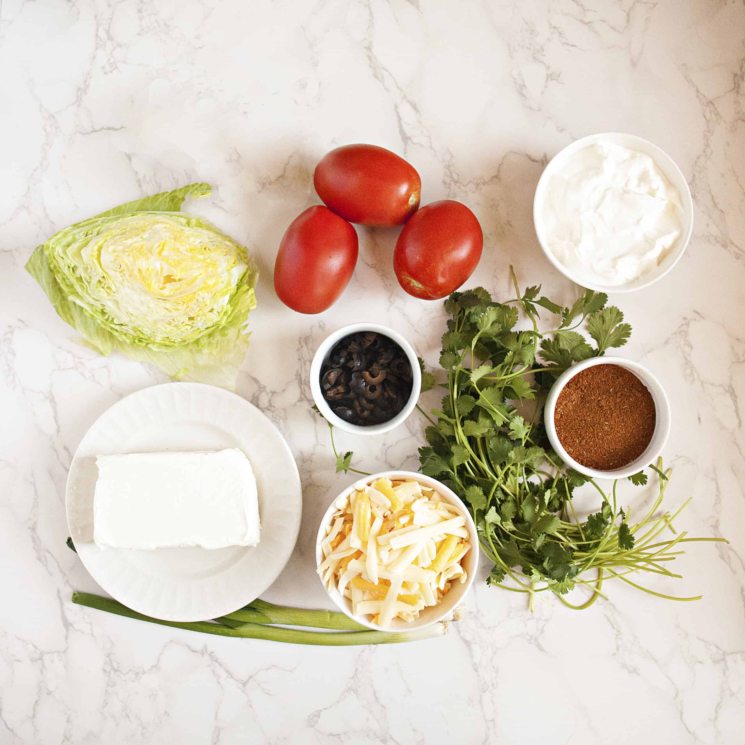 Ingredients for quick cream cheese taco dip: Cream cheese, sour cream, taco seasoning, lettuce, roma tomatoes, sliced black olives, cilantro, green onions, and shredded cheese, on a white marble background.