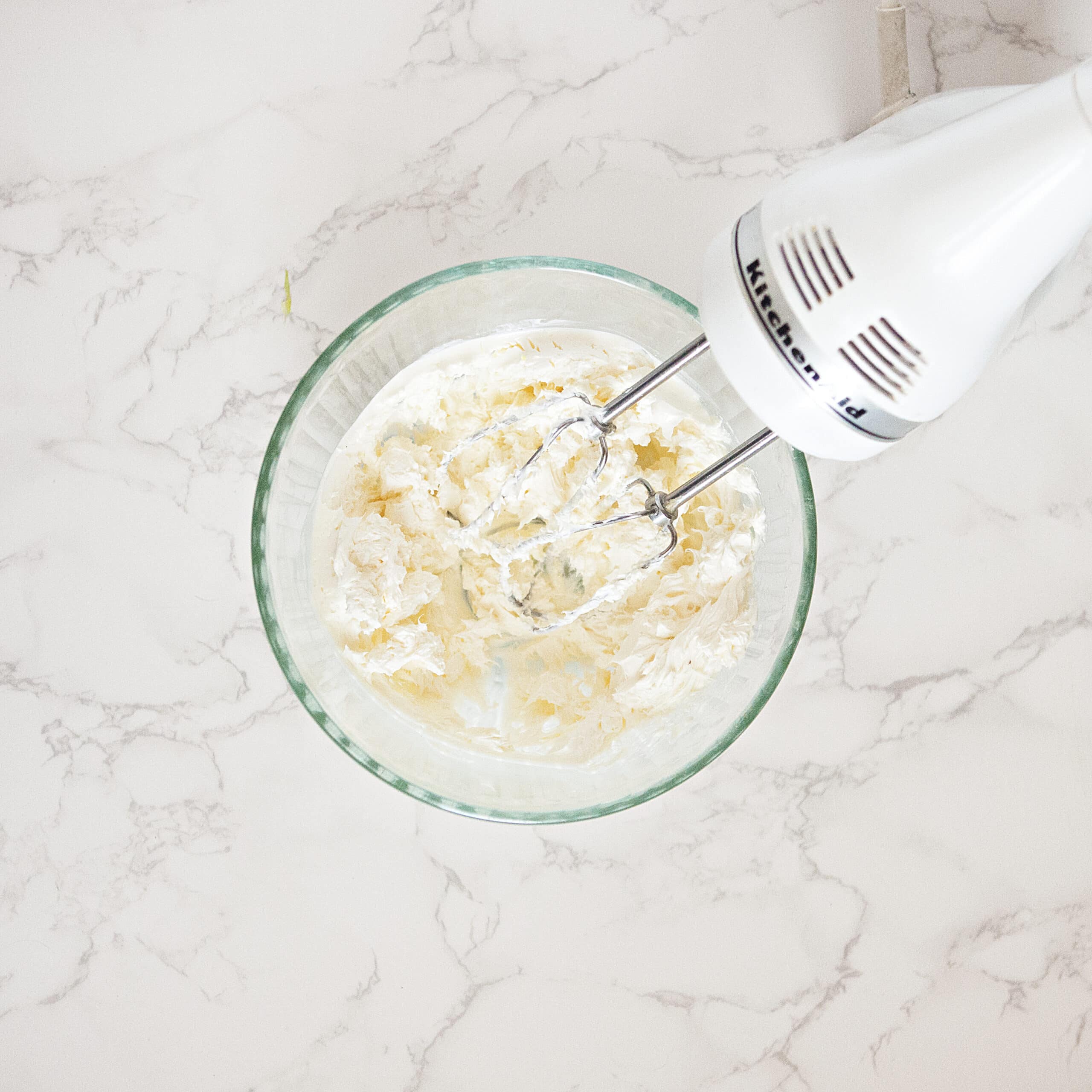 A glass bowl containing whipped cream cheese next to an electric hand mixer on a white marble background.