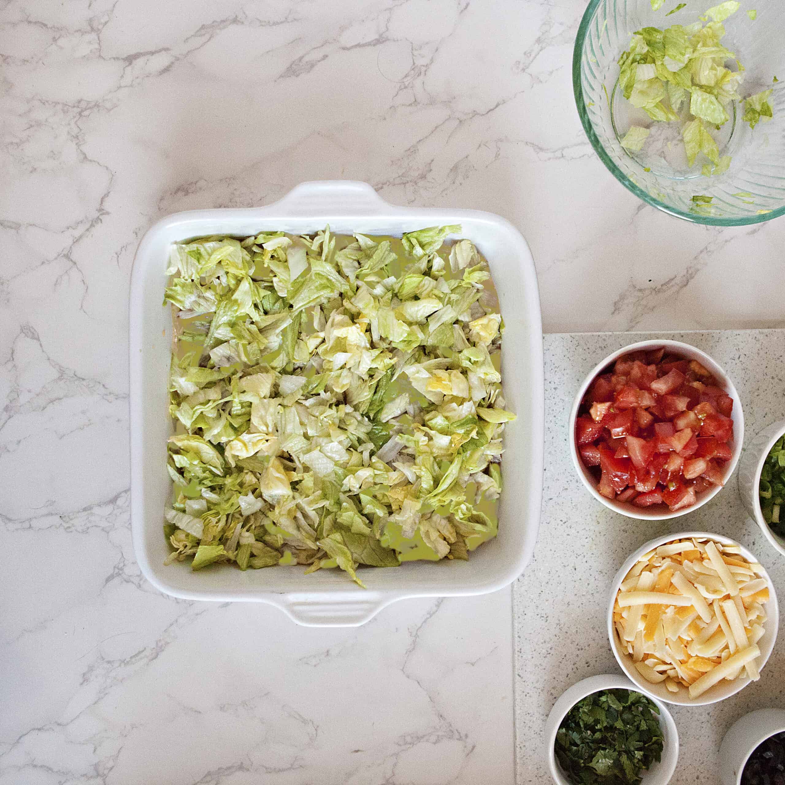 A white square baking dish containing a light orange dip covered with chopped lettuce, alongside more toppings in small bowls, on a white marble background.